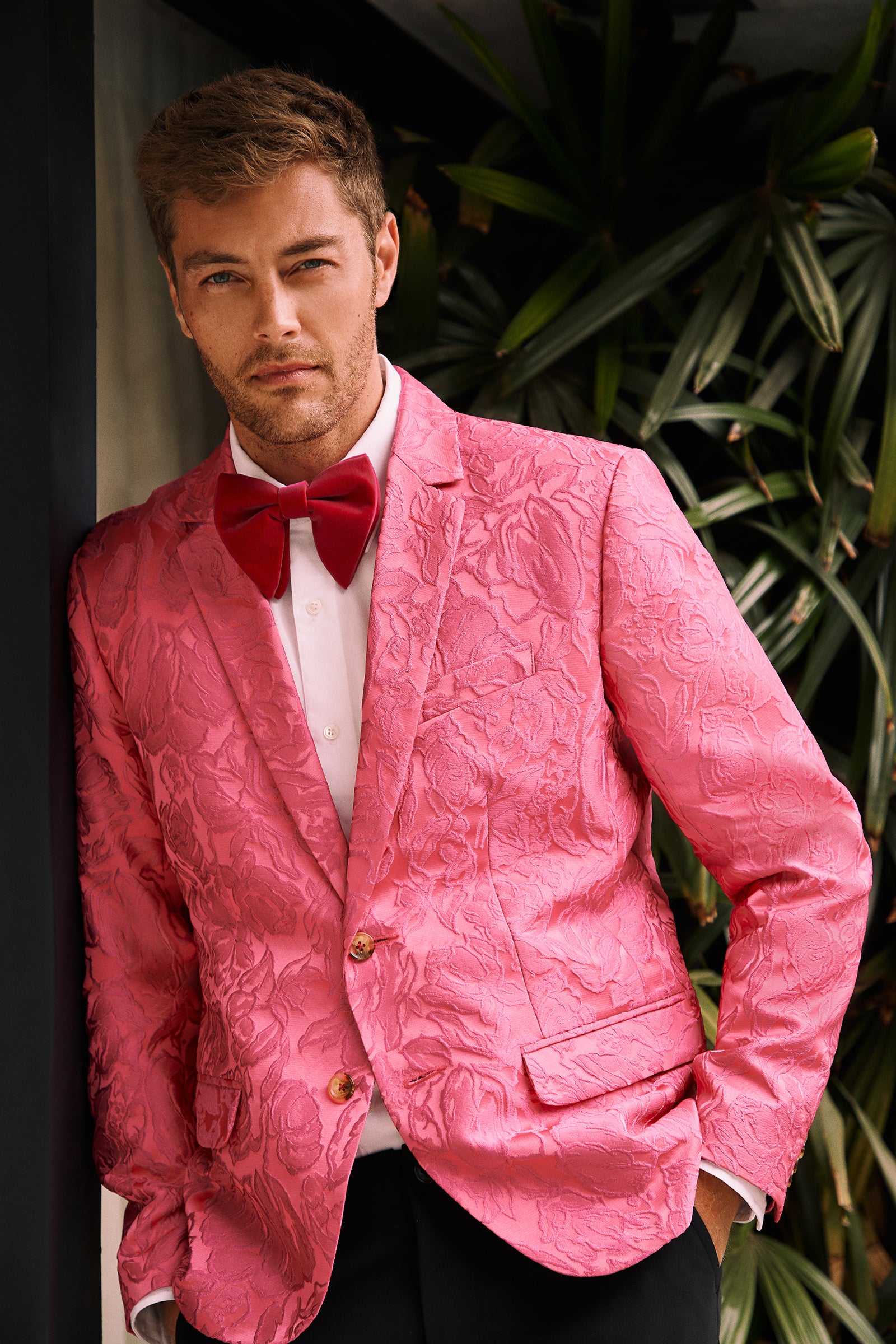 A man wearing the Thurston Blazer - Sweetheart Jacquard, paired with a white shirt and red bow tie, stands with one hand in his pocket, leaning against a wall adorned with green leafy plants.