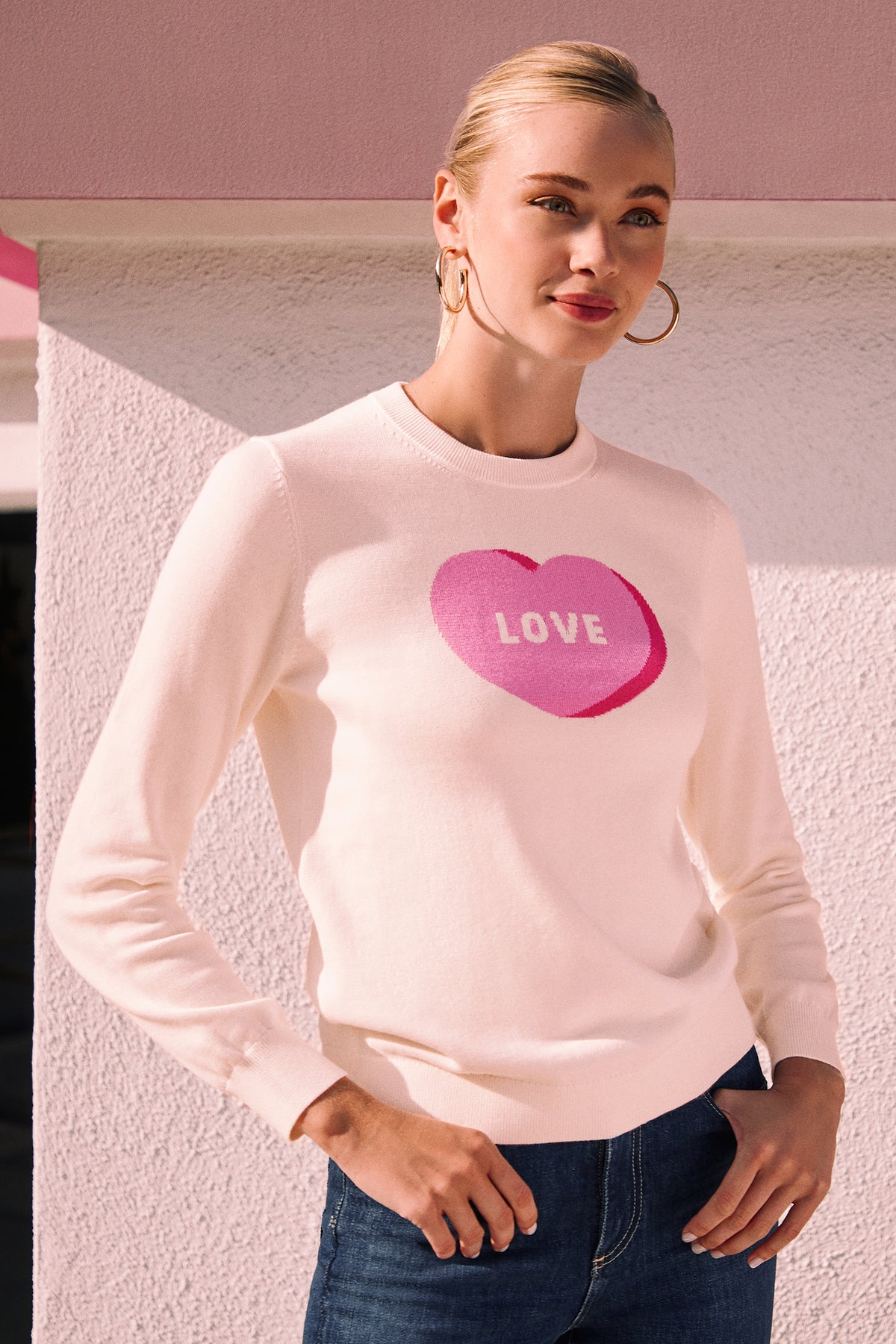 A woman with blonde hair in a bun wears hoop earrings, blue jeans, and the Love Merino Wool Crewneck Sweater, which is white with a pink heart and the word LOVE. She stands before a pastel pink and white wall, smiling gently.