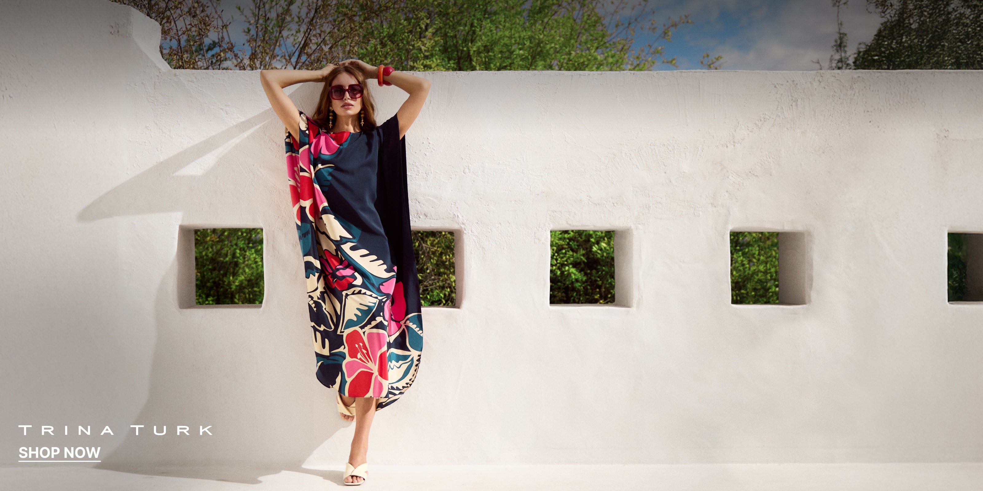 Woman wearing a colorful dress standing against a white wall with a blue sky and trees in the background, Trina Turk brand visible.