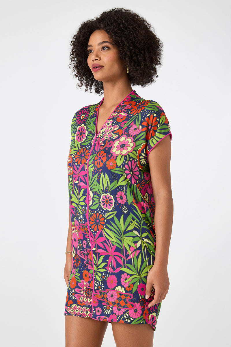 A woman with curly hair wears the Tarashi Short-Sleeve Mini Dress - Fumi Floral, showcasing vibrant pink, green, orange, and purple flowers and leaves, standing against a plain white background.