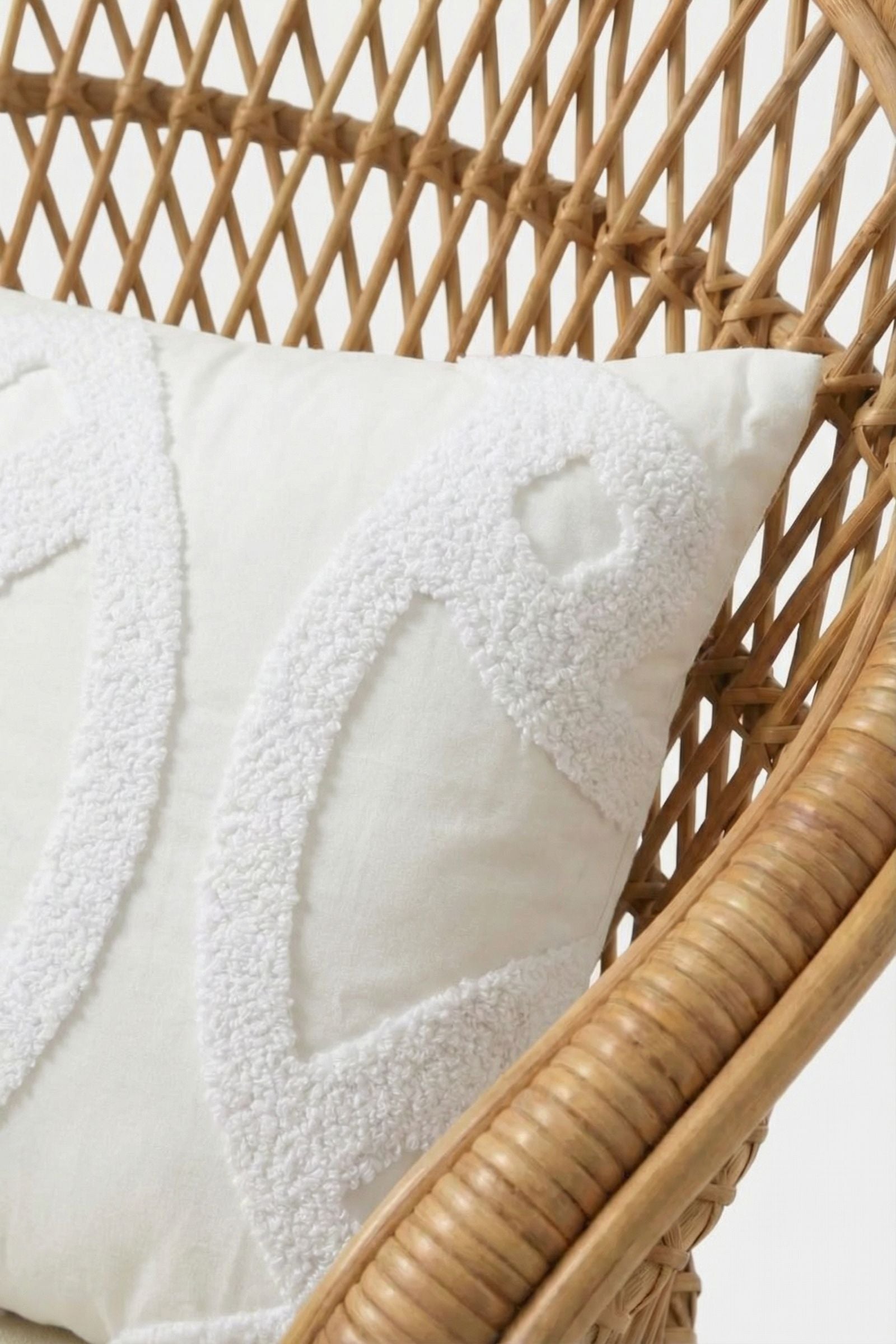 A close-up of the Santorini White Tufted Throw Pillow 12"X24", featuring abstract patterns on white fabric, resting on a woven rattan chair against a plain white background.