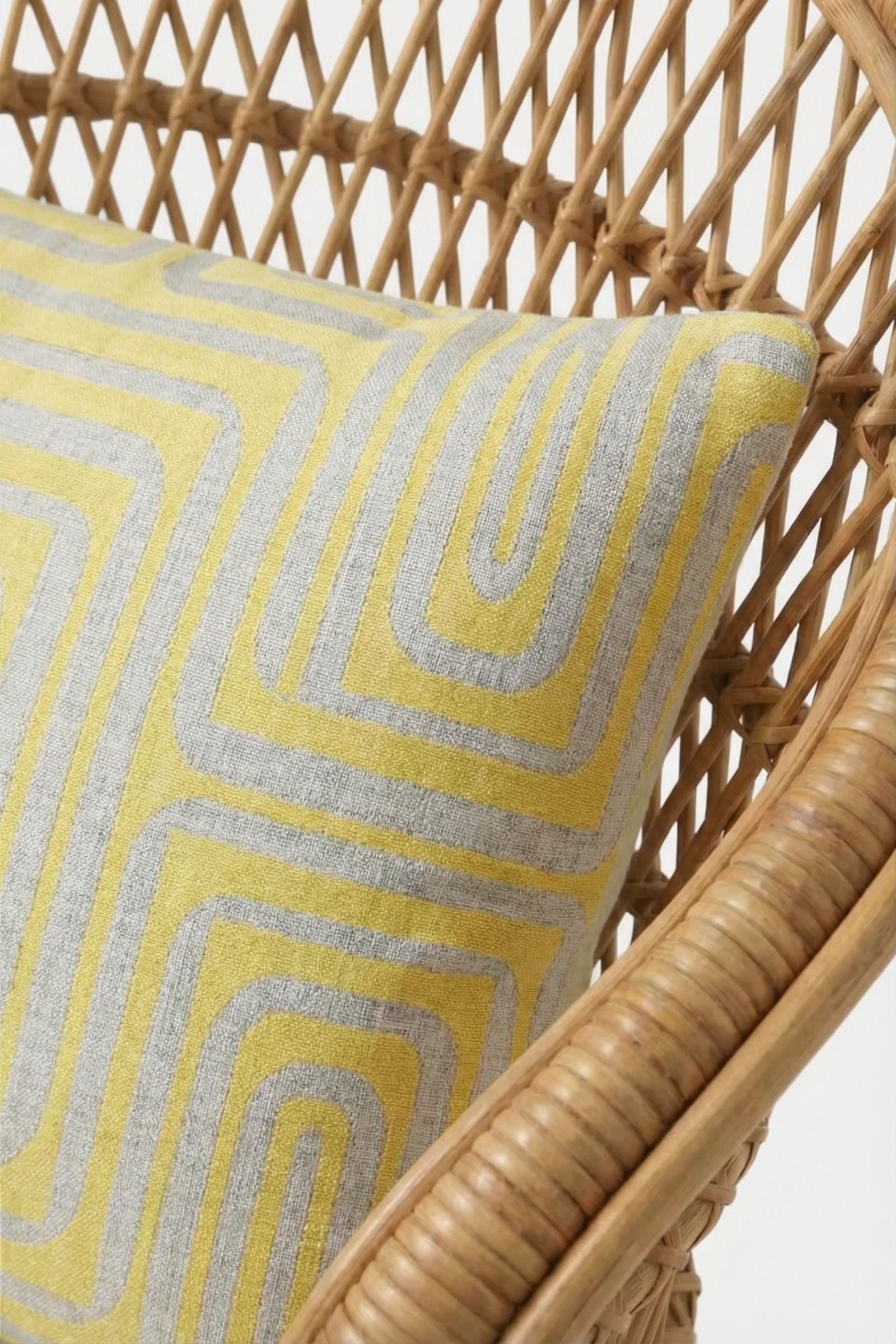 A close-up of a rattan chair with the Amazing Maze Embroidered Pillow, featuring a geometric yellow and gray pattern.
