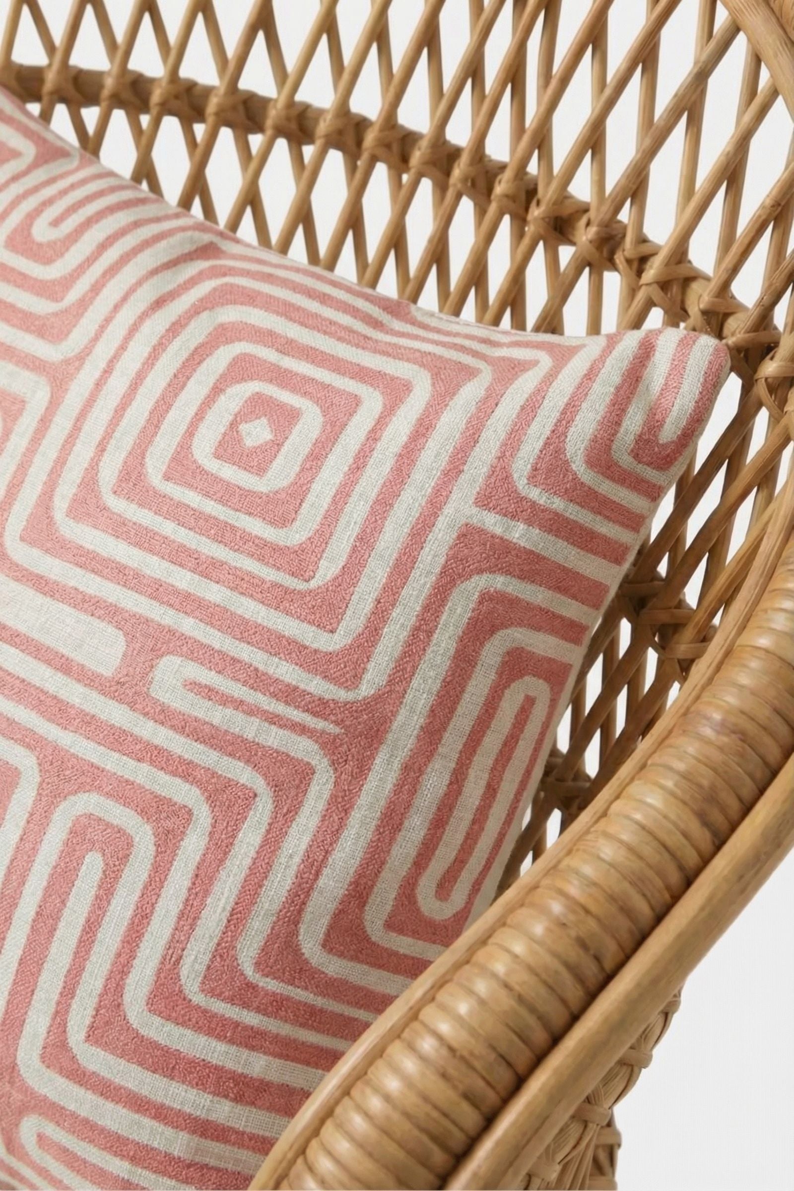 A close-up of a wicker chair with the Amazing Maze Embroidered Pillow, showcasing its pink and white geometric square and maze-like designs.