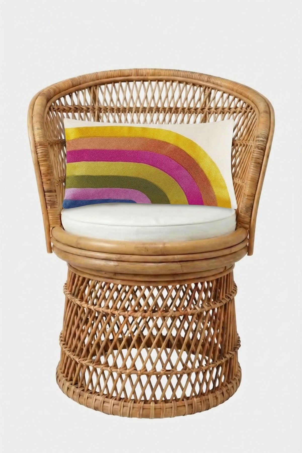 A wicker chair with a round white cushion features the Calistoga Embroidered Rainbow Lumbar Pillow, showcasing yellow, pink, green, and blue embroidery on a white background. The chair is set against a plain white backdrop.