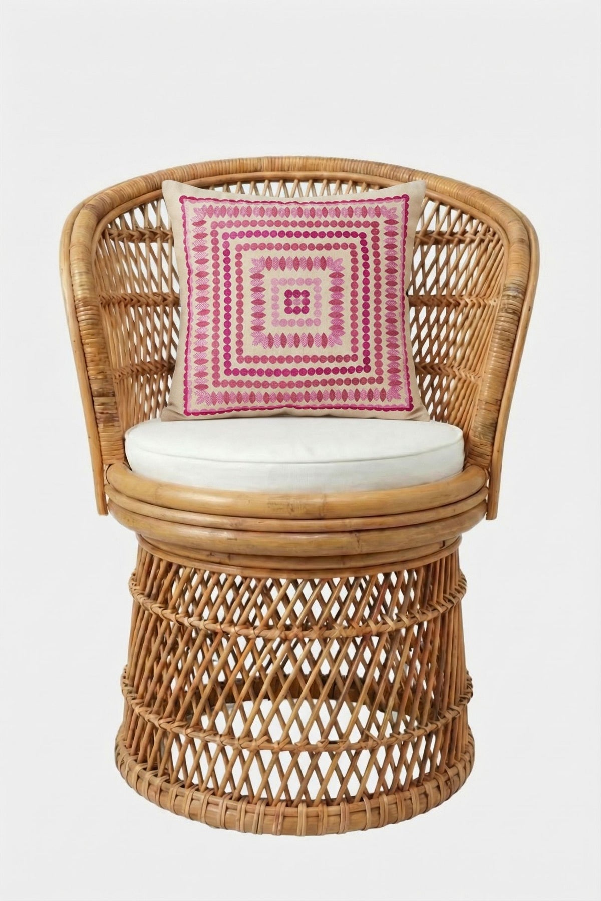 A round wicker chair with a white cushion and the Carmel Embroidered Pillow 20x20", featuring a pink geometric pattern, set against a plain white background.