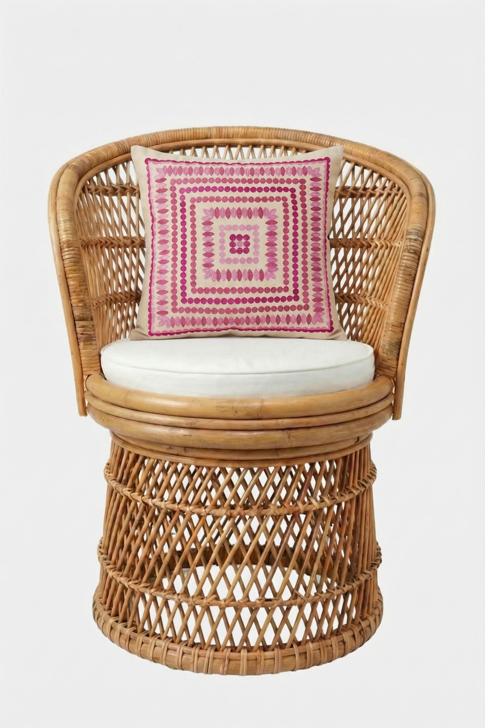 A round wicker chair with a white cushion and the Carmel Embroidered Pillow 20x20", featuring a pink geometric pattern, set against a plain white background.