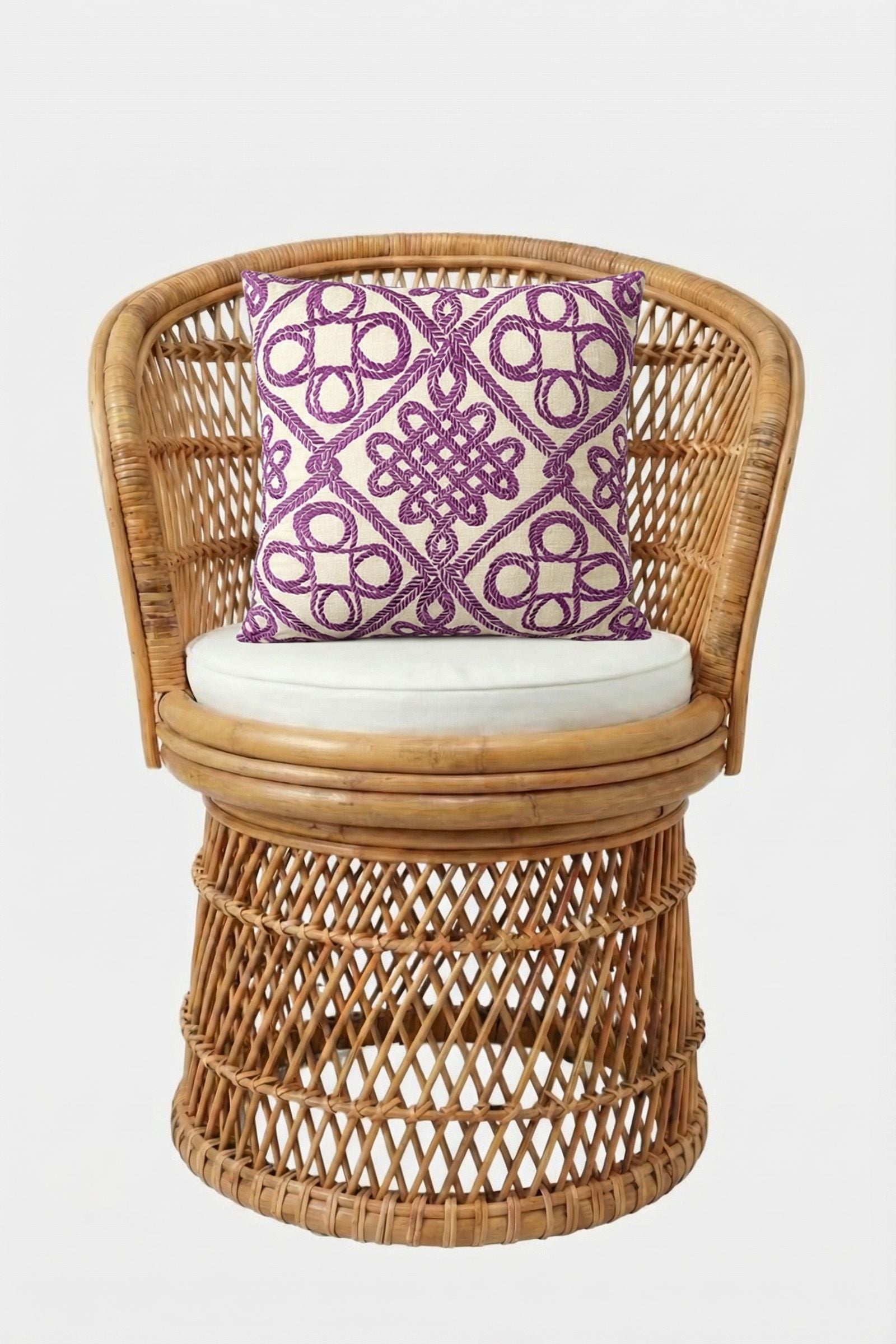 A round wicker chair with a white cushion and the Costa Mesa Embroidered Throw Pillow, showcasing a purple geometric design, set against a plain white background.