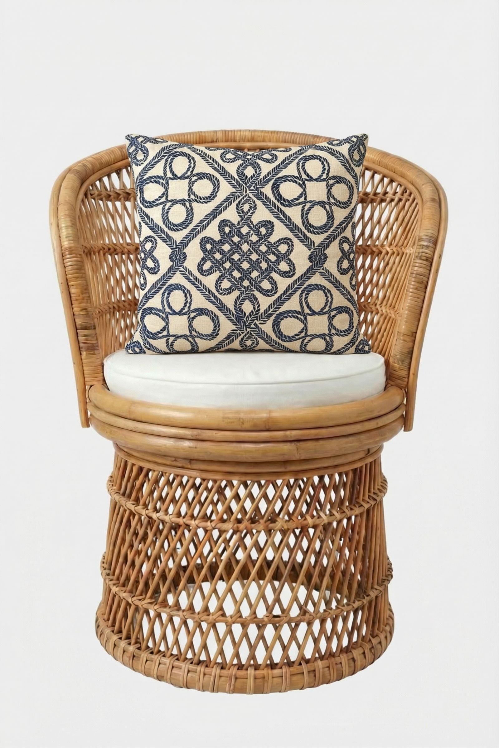 A rattan chair with a rounded backrest and white seat cushion, featuring the Costa Mesa Embroidered Throw Pillow—a decorative square pillow with a navy blue geometric pattern on a beige background.