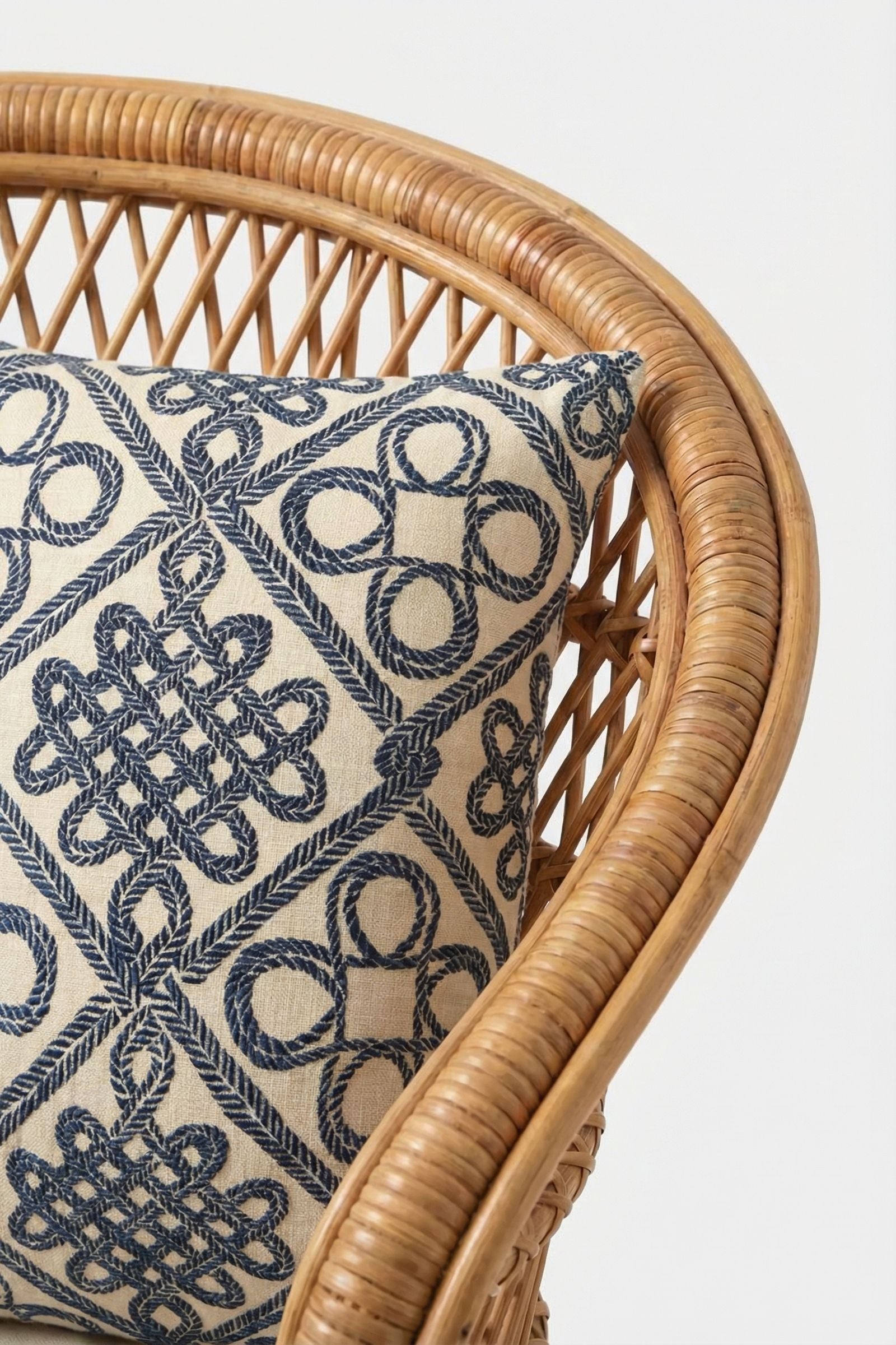 A close-up of the Costa Mesa Embroidered Throw Pillow with an intricate blue geometric and knot pattern on a cream cushion, set on a rattan chair against a plain white background.