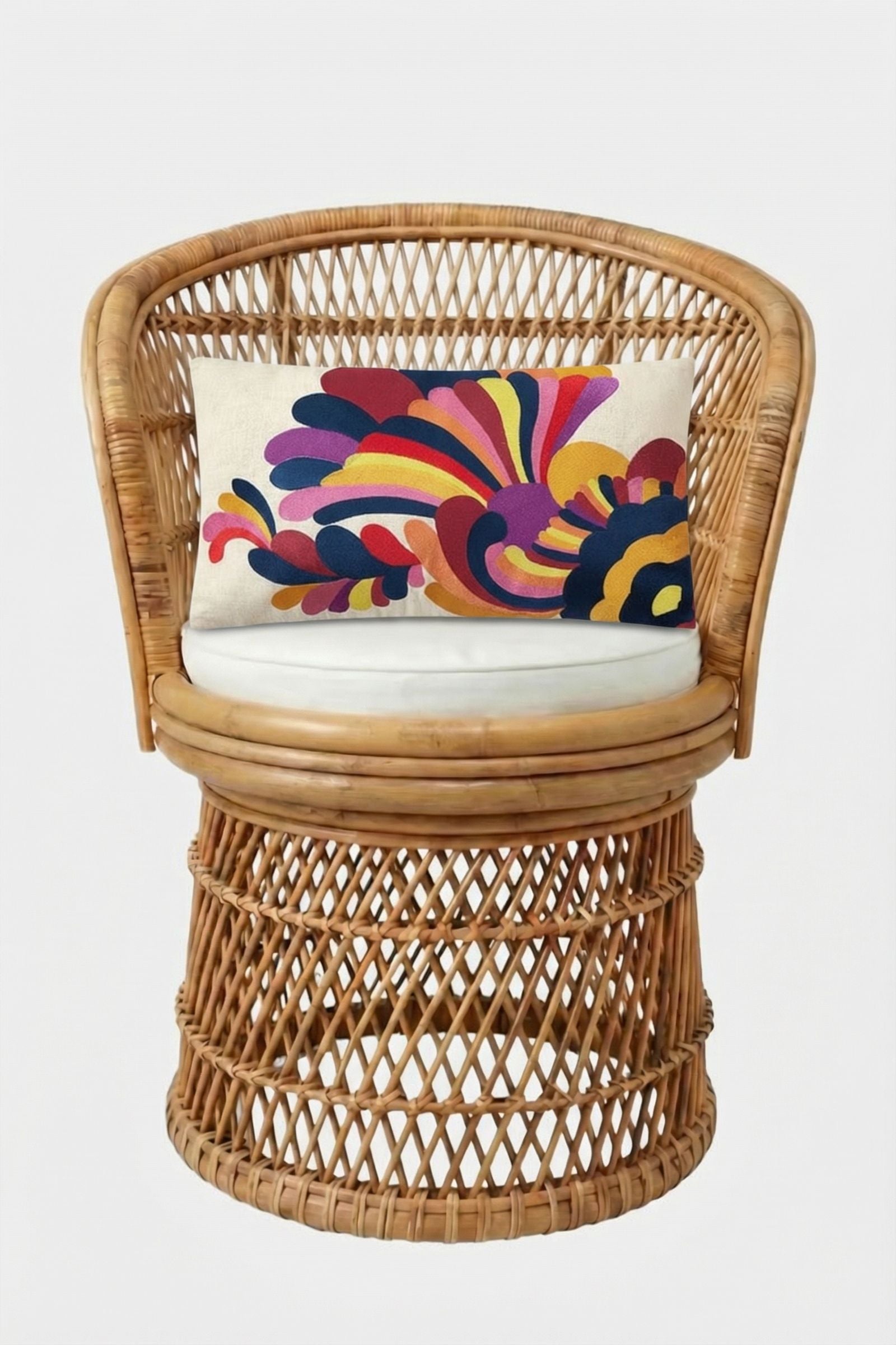 A round wicker chair with a white cushion and the Escondido Embroidered Lumbar Pillow, showcasing a colorful abstract floral design in red, blue, yellow, and purple on white, set against a plain white background.