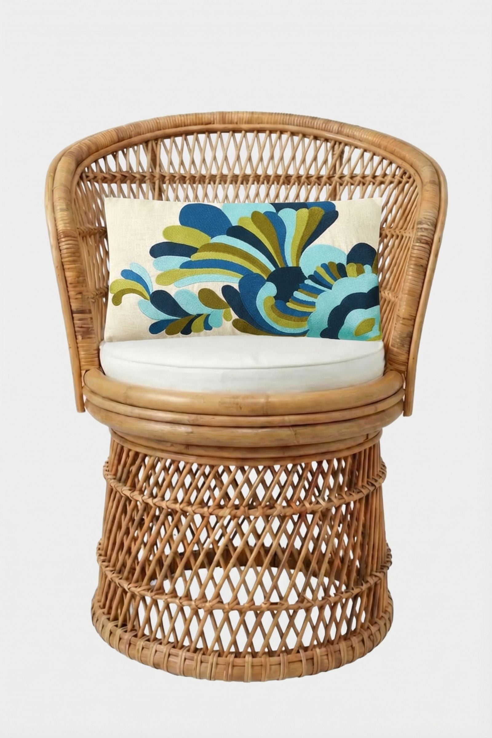 A round wicker chair with a white cushion and the Escondido Embroidered Lumbar Pillow, displaying a blue, green, and yellow abstract pattern, set against a plain white background.