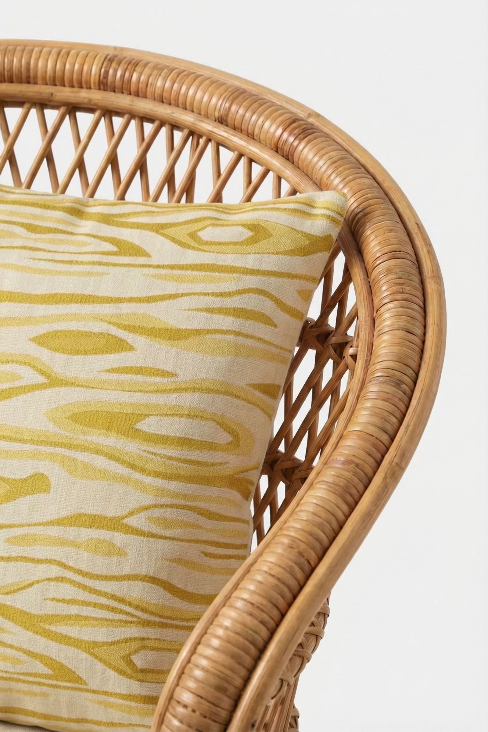 A close-up of a wicker chair with a curved backrest, featuring the Monterey Embroidered Throw Pillow in yellow and cream with a woodgrain-inspired pattern.