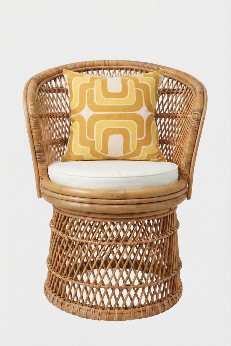 A rattan chair with a round white cushion and an Ogee Amber Embroidered Square Pillow showcasing a geometric pattern, set against a plain white background.