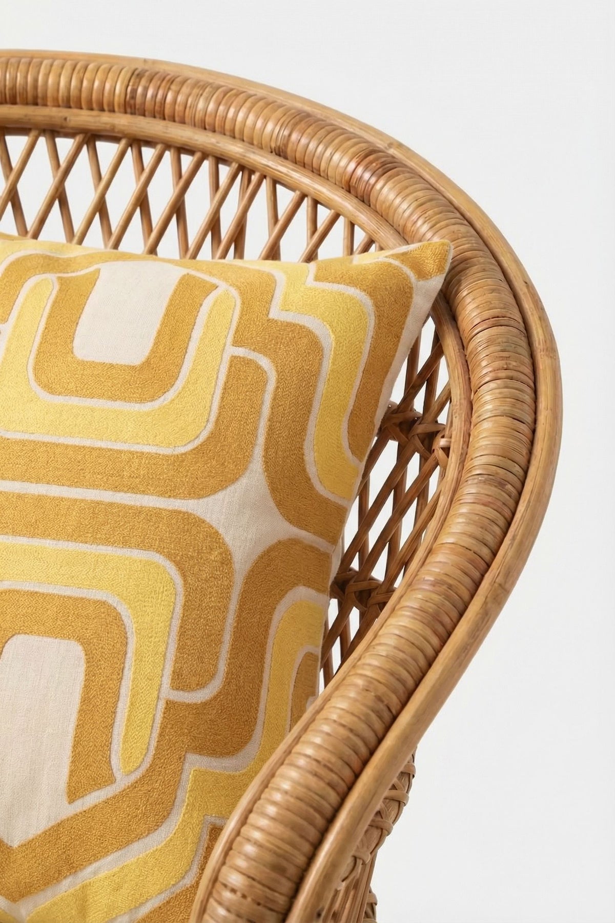 Close-up of a wicker chair with the Ogee Amber Embroidered Square Pillow, showcasing a yellow and white geometric, retro-inspired design. The background is plain white.