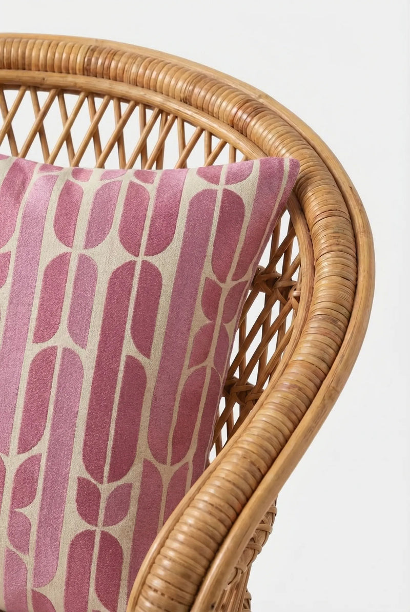 A close-up of the Palmdale Emb Pillow 20X20", a decorative throw pillow with a geometric pink and beige pattern, placed on a wicker chair against a plain white background.