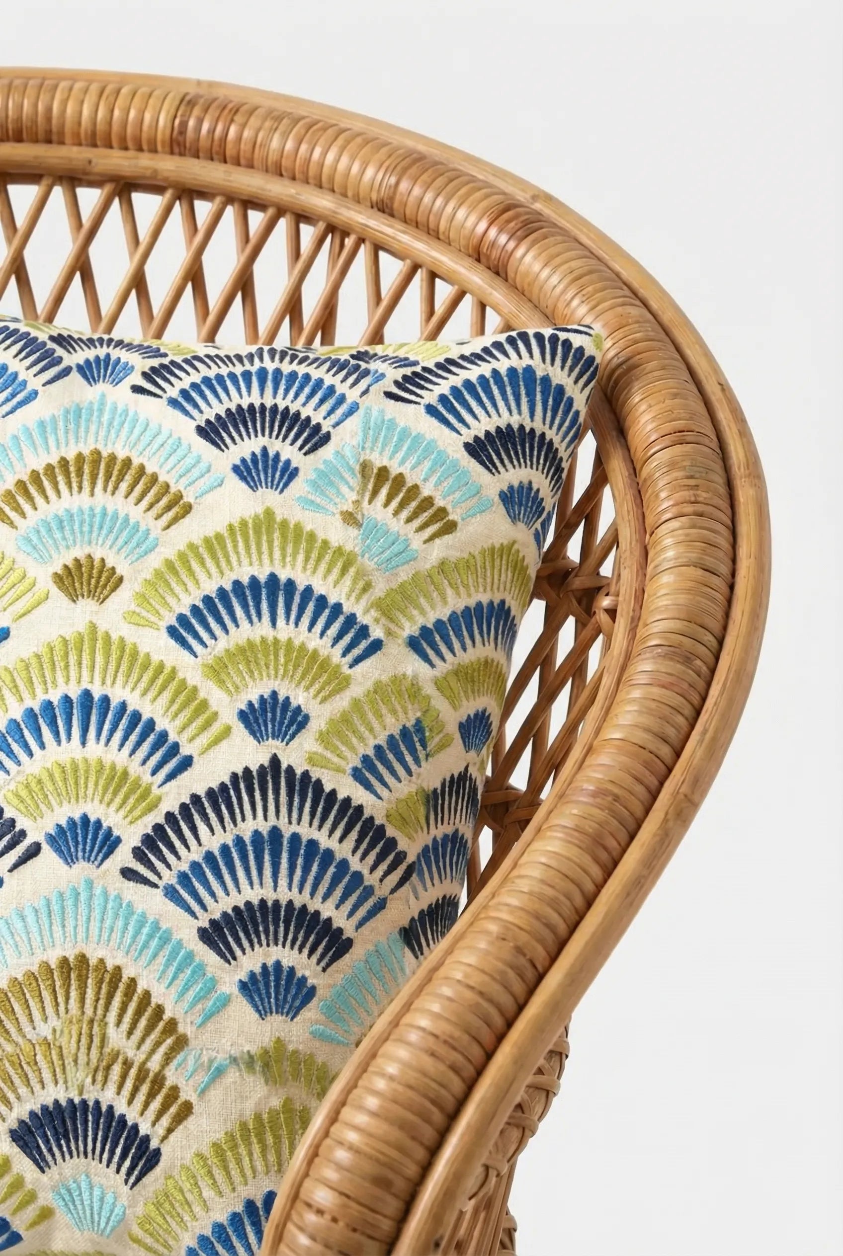 A close-up of a curved wicker chair with the Pasadena Embroidered Euro Throw Pillow, showcasing a blue, green, and white geometric fan pattern. The plain white background highlights the decorative pillow.