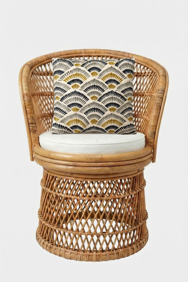 A round rattan chair with an intricate woven pattern and a white seat cushion, styled with the Pasadena Embroidered Square Pillow, which features a geometric fan-like design in black, white, and yellow. The background is plain white.