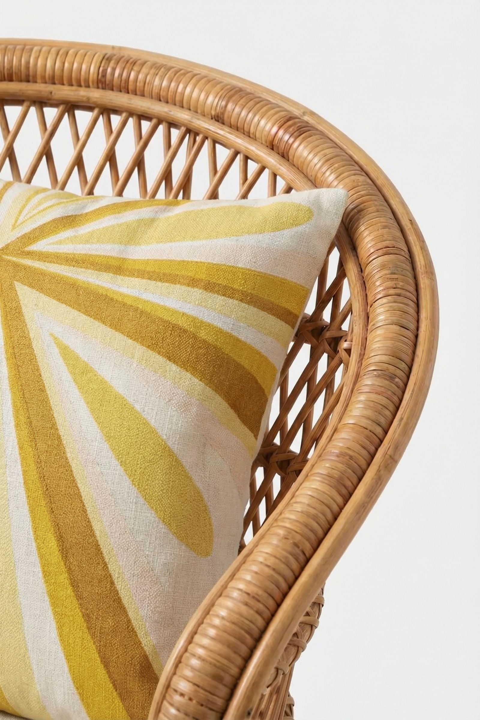 A close-up of the Shante Embroidered Pillow, showcasing its yellow and white abstract, radiating pattern on a rattan chair with a woven curved back and armrest against a plain white background.