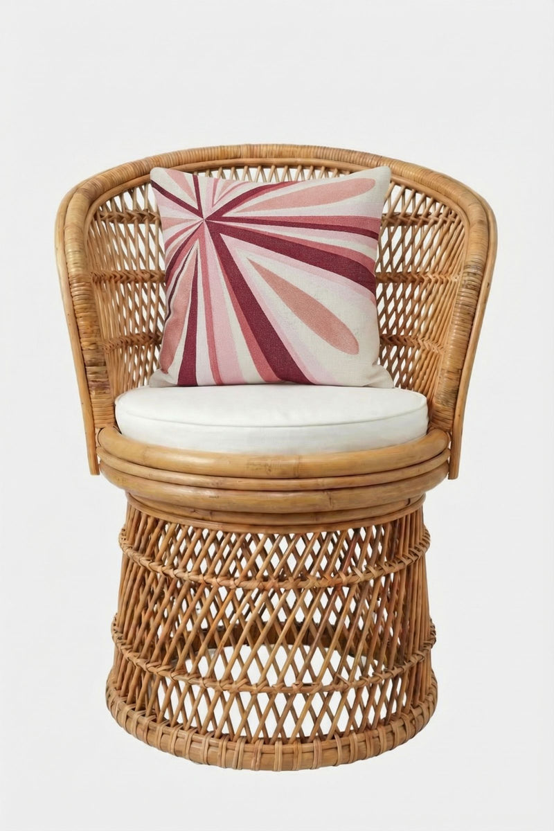 A round rattan chair with an open-weave design, white seat cushion, and the Shante Embroidered Square Pillow featuring bold pink and red geometric lines on white, set against a plain white backdrop.