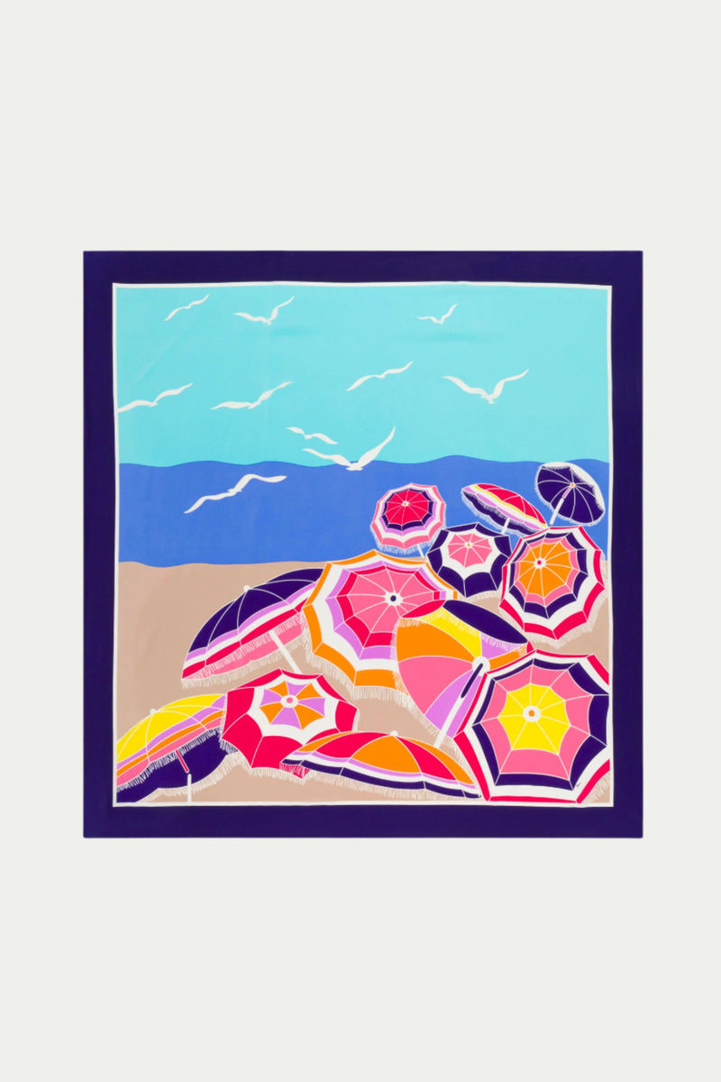 The Umbrella Scarf - Parapluie Silk features a colorful beach scene with vibrant pink, red, orange, and purple umbrellas on sandy shore, blue sea and sky, white birds flying, all bordered in dark blue. Color: Multi.