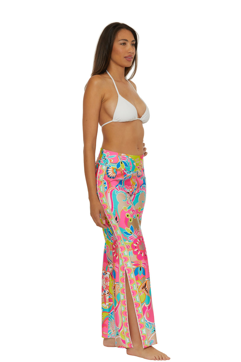 A woman stands barefoot in profile against a plain white background, wearing the Venus Swim Cover-Up Pant—bright, multicolored with a floral pattern and side slits—paired with a white bikini top.