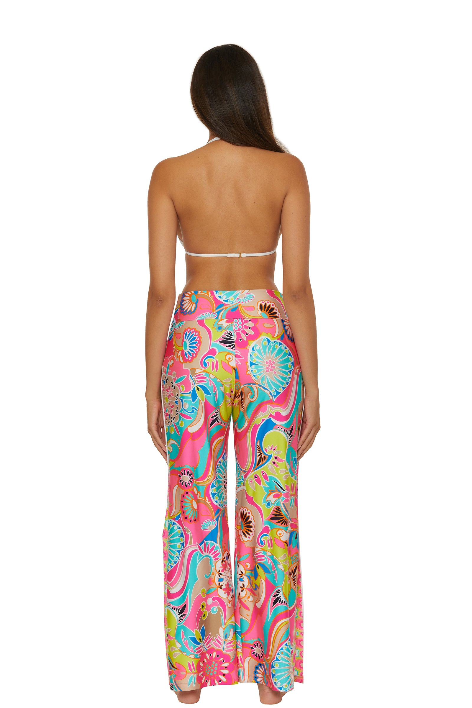 A woman stands with her back to the camera, wearing a white halter bikini top paired with the Swim Cover-Up Pant - Venus—high-waisted, wide-leg pants featuring a colorful, abstract floral print.
