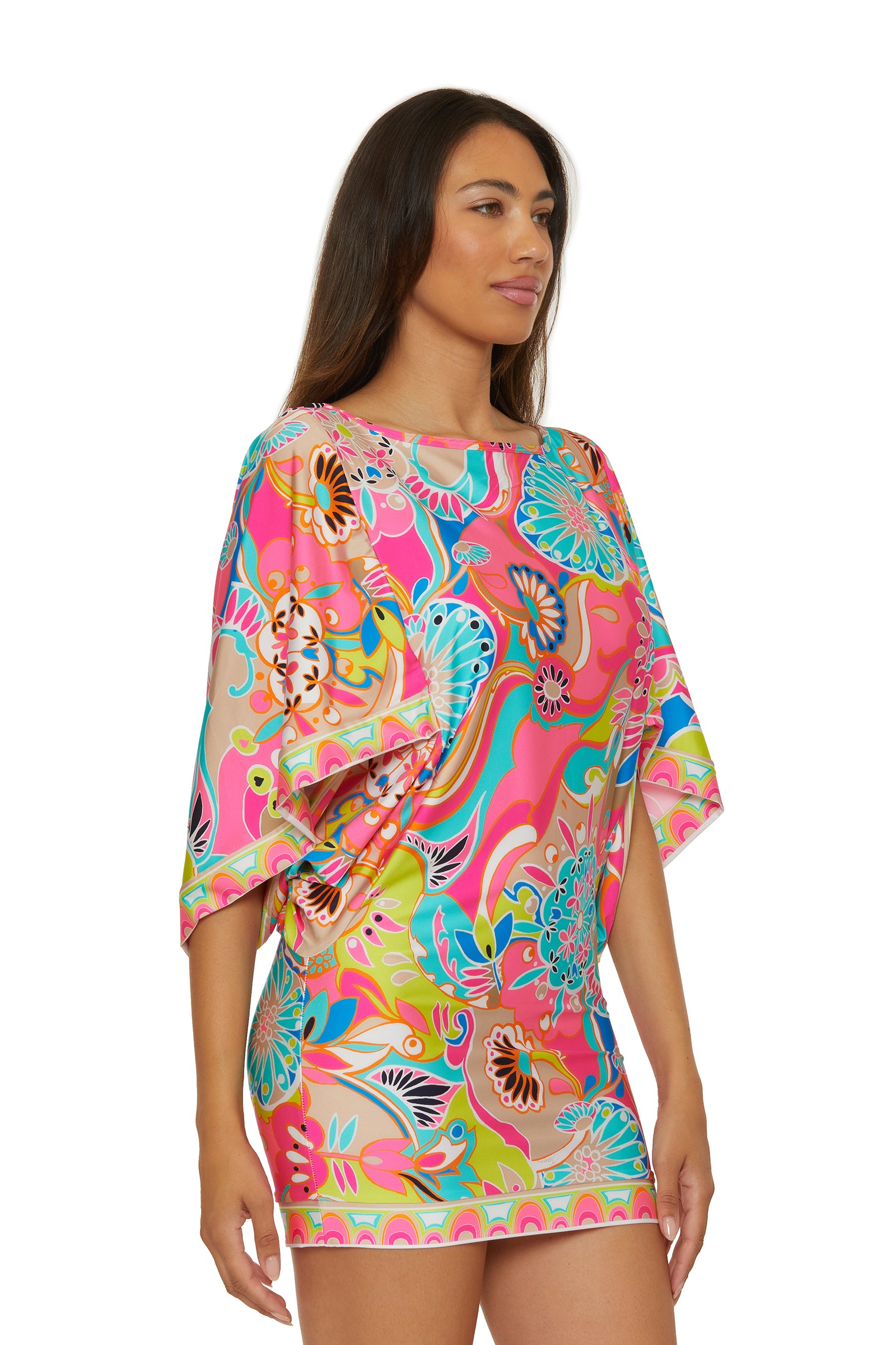 A woman models the Casablanca Tunic Cover-Up by Venus, a vibrant floral mini dress with loose sleeves in pink, blue, yellow, and green. She stands against a white background, looking to the side and smiling.