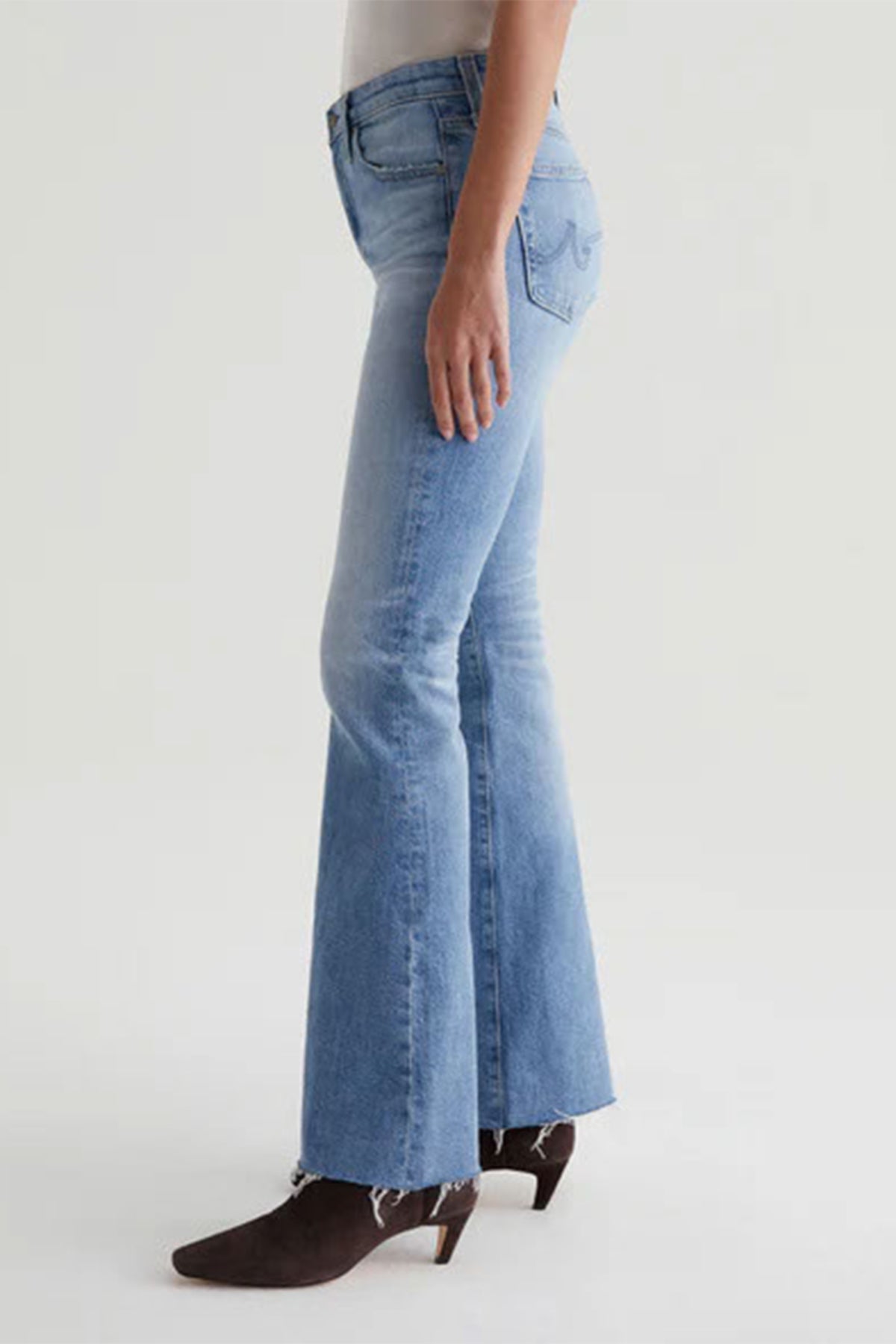 A person wearing AG Womens Farrah Bootcut Blue Jeans—a light wash, high-rise bootcut style with a raw hem—and brown heeled ankle boots, shown from the side against a plain light background.