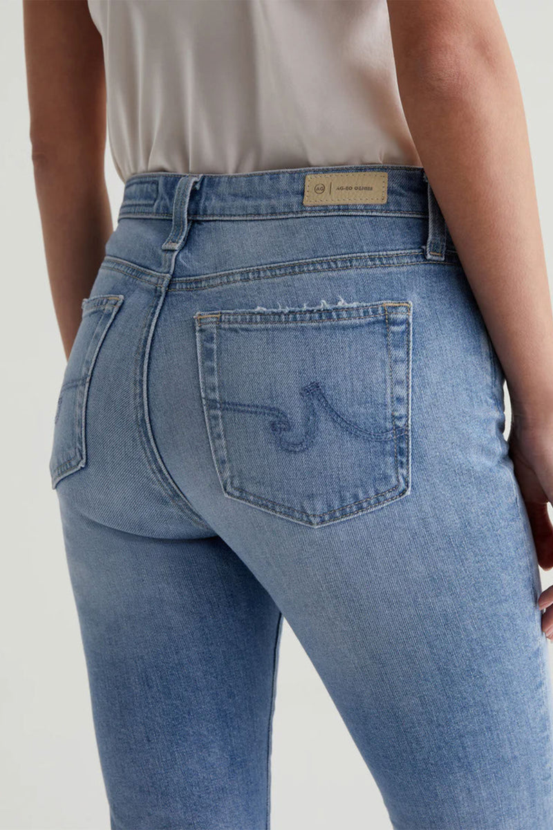 The image shows a person from the waist down wearing AG Womens Farrah Bootcut Blue Jean, light wash blue with a visible AG-ed Denim label above the right back pocket, paired with a white top.