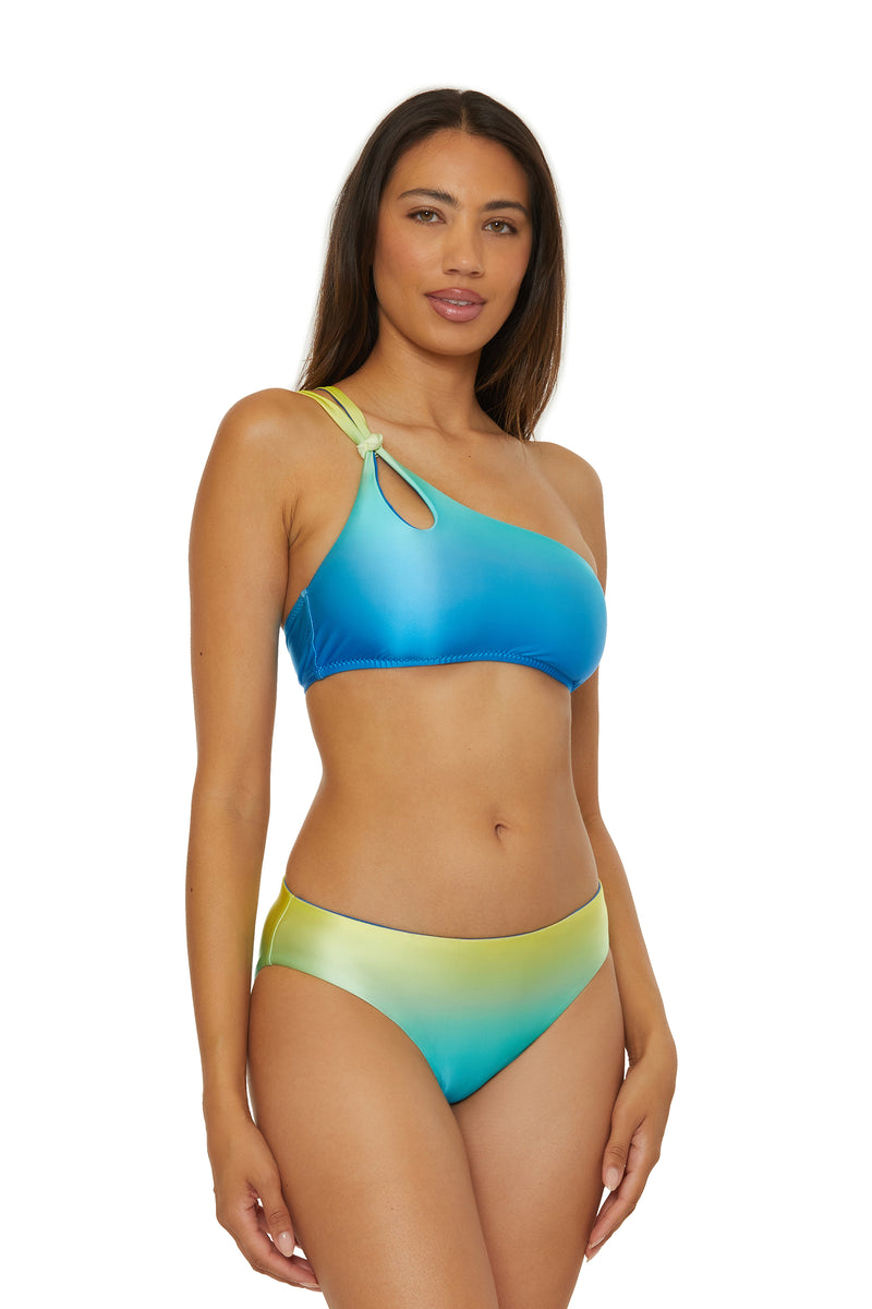 A woman models the One Shoulder Swim Top - West Palm, featuring a blue-to-yellow gradient, knot and keyhole strap detail. She has long straight hair and poses against a plain white background.