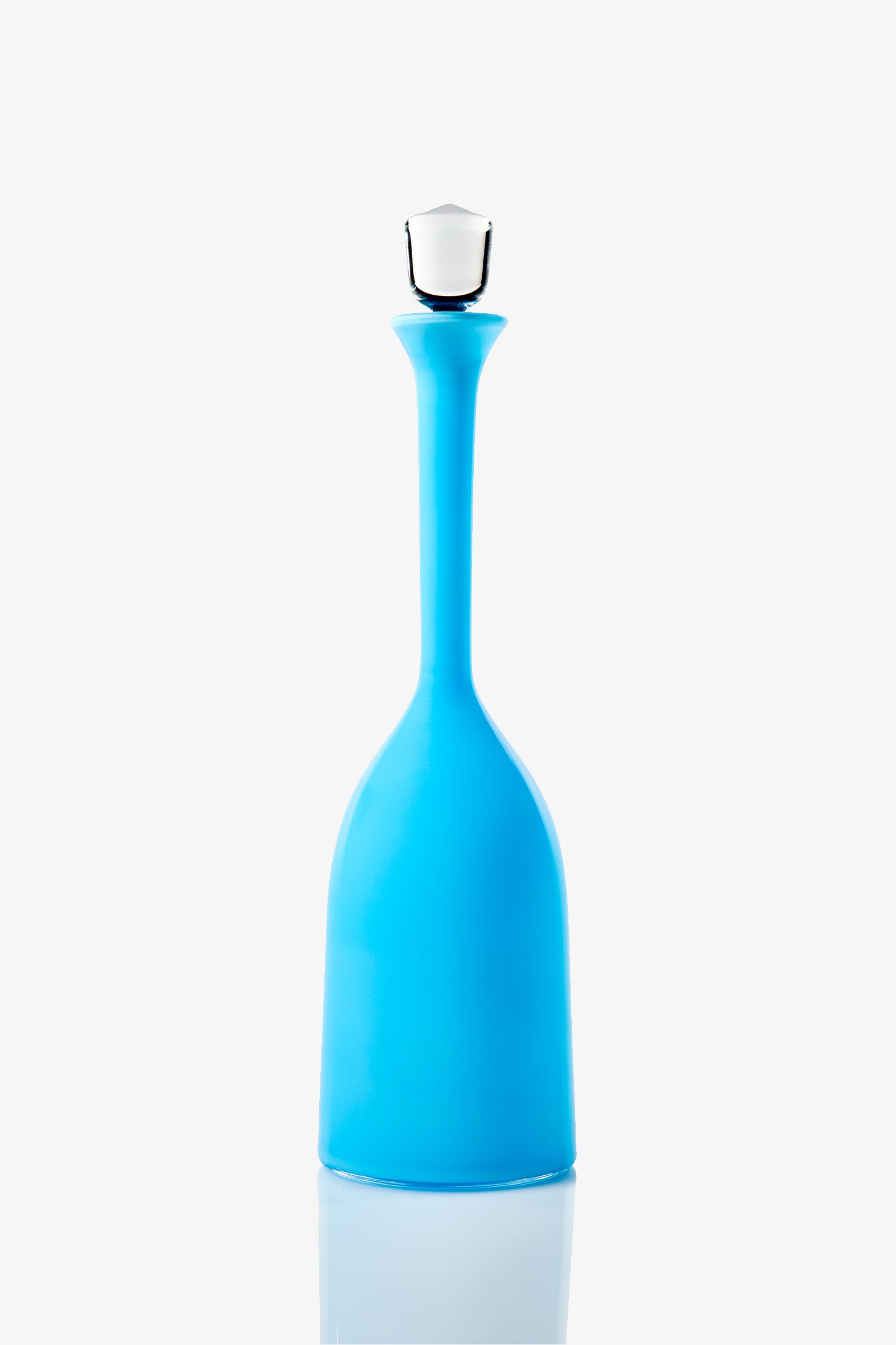 The Wexler Wide Bottle, featuring a tall, slender blue glass body with a narrow neck and a white stopper, stands upright on a white reflective surface against a plain white background.