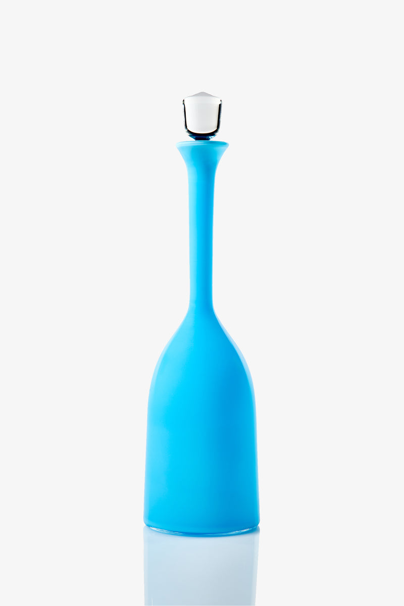 The Wexler Wide Bottle, featuring a tall, slender blue glass body with a narrow neck and a white stopper, stands upright on a white reflective surface against a plain white background.