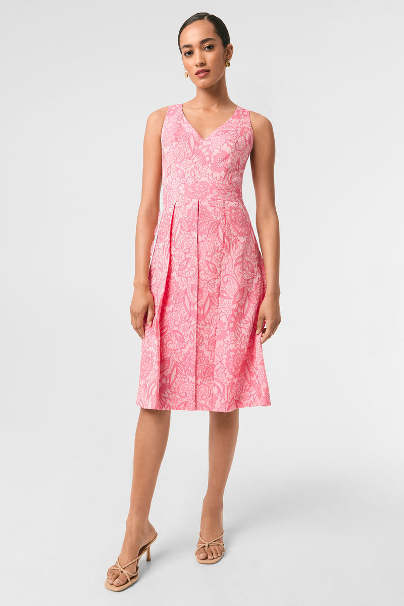 A woman poses against a plain background in the Jayme Sleeveless Dress - Jolie Jacquard in Eden Roc Rose, featuring a V-neckline, knee-length pleated skirt, and nude heeled sandals.