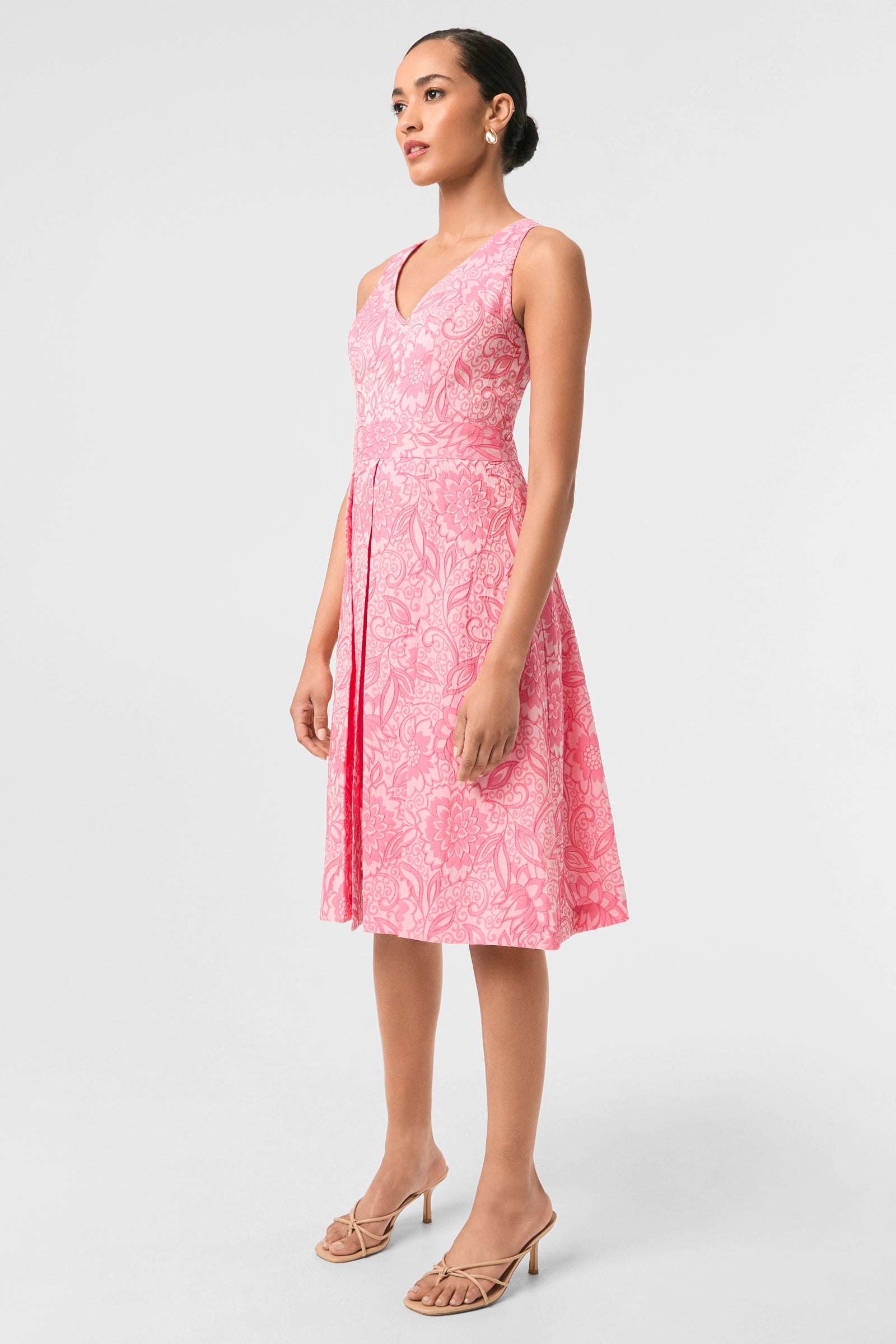 Woman wearing the Jayme Sleeveless Dress - Jolie Jacquard, a knee-length pink floral V-neck dress, paired with nude heeled sandals, standing against a plain light background.