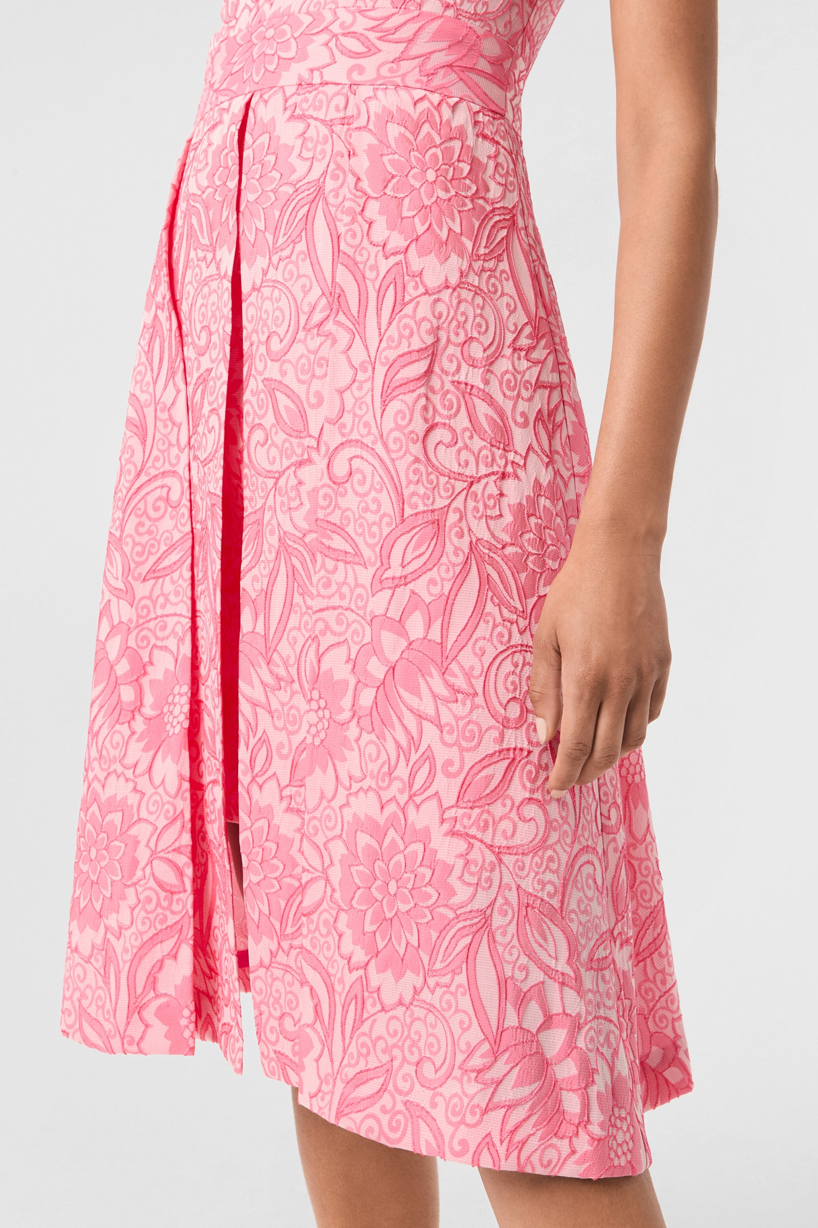Someone wearing the Jayme Sleeveless Dress - Jolie Jacquard, a pink and white knee-length dress with a floral and leaf pattern, shows their right arm and part of their torso against a plain light background.
