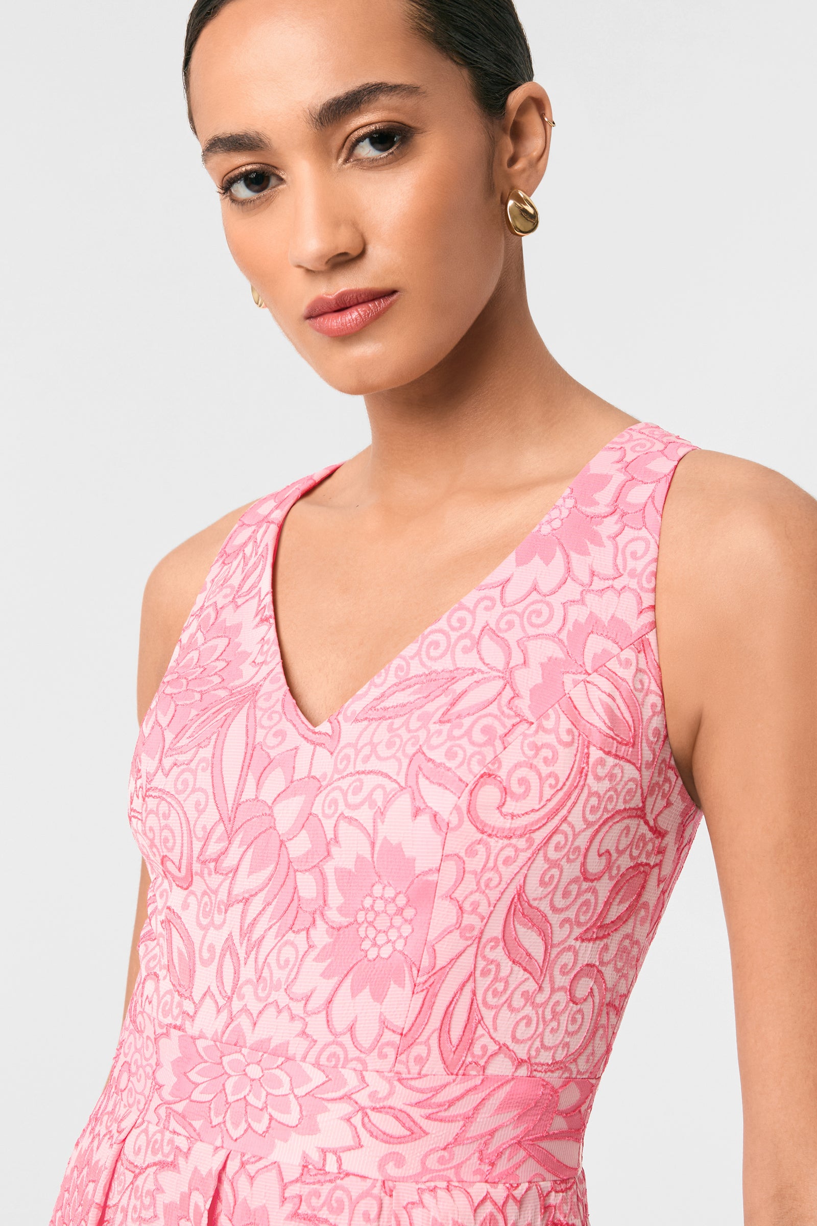 A woman with neatly styled hair and a calm expression wears the Jayme Sleeveless Dress - Jolie Jacquard, featuring a pink floral pattern and V-neck, paired with gold hoop earrings, standing against a plain light background.