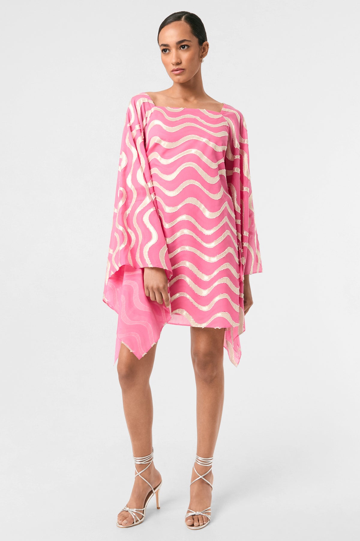 A woman poses against a light background in the Aqualane Side Flounce Dress - Pinku, featuring long sleeves, white wavy patterns, an asymmetrical hem, and paired with white strappy heels. Her hair is pulled back.