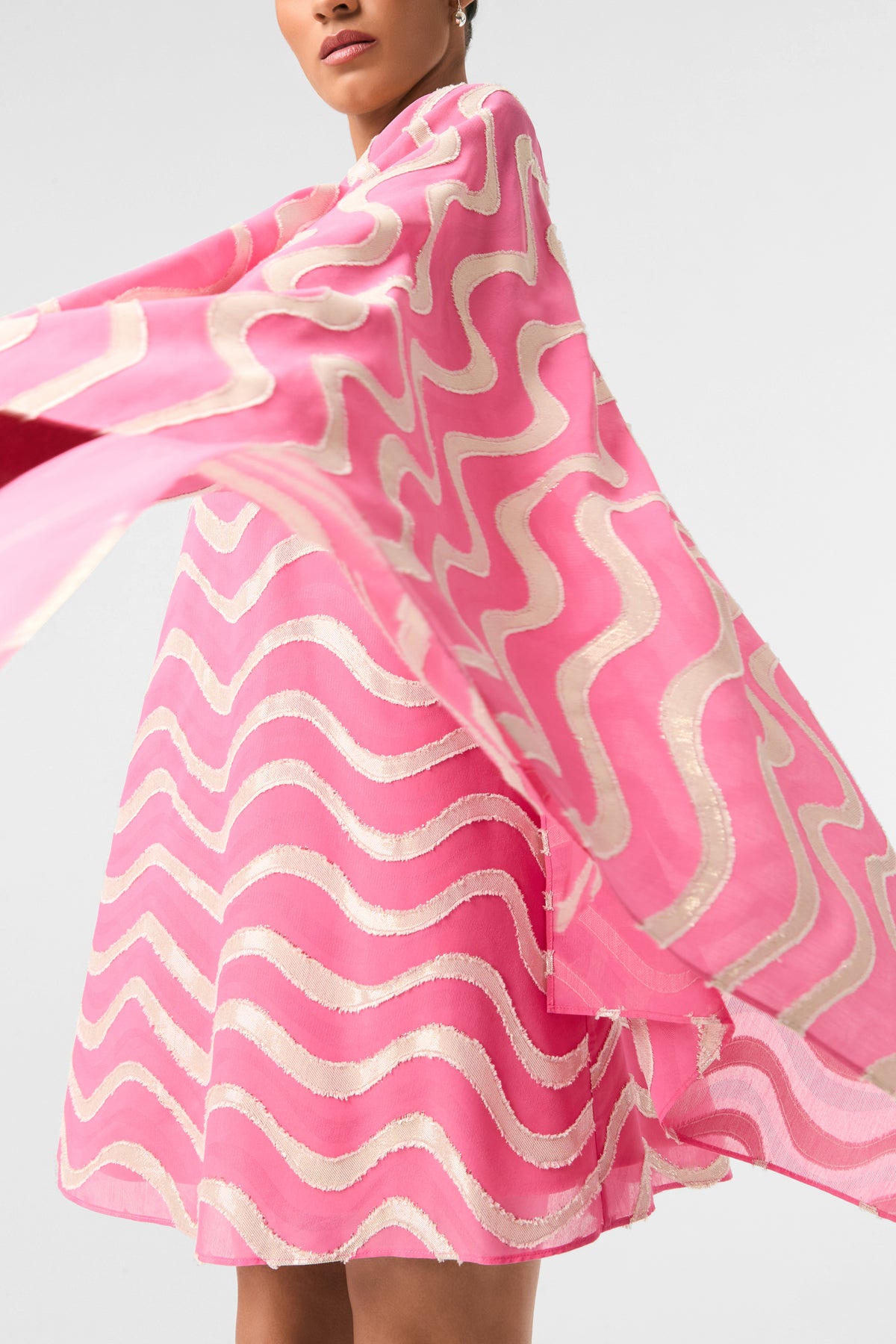 A person wearing the Aqualane Side Flounce Dress - Pinku, a flowing bright pink dress with wavy gold and white patterns. The fabric flares outward, creating a sense of movement against a plain, light background.