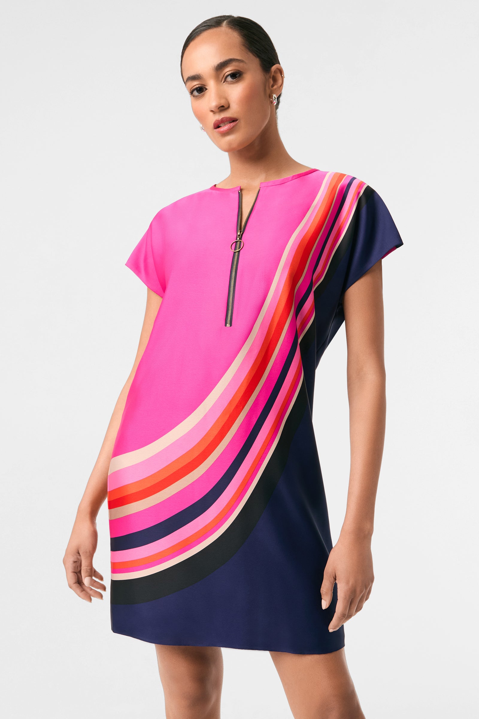 A woman wears the Hasil 2 Short-Sleeve Shift Dress, featuring bold diagonal stripes in pink, orange, red, and black on a pink and navy background. The dress is knee-length with a front zipper; she has her hair pulled back.