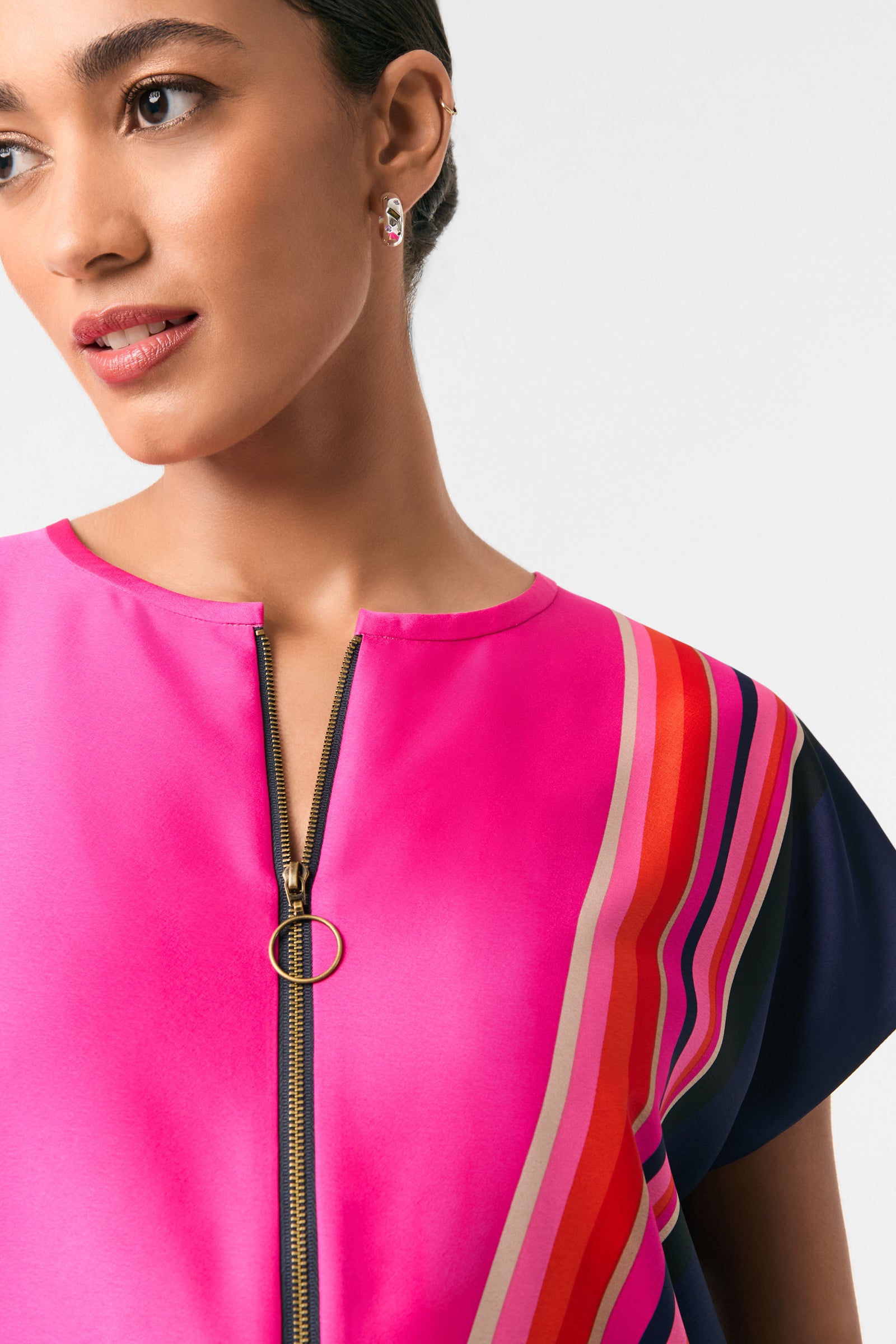 A woman models the Hasil 2 Short-Sleeve Shift Dress, a bright pink dress with a front zipper and colorful diagonal stripes, gazing to the side. Only her upper face and torso appear against a plain white background.