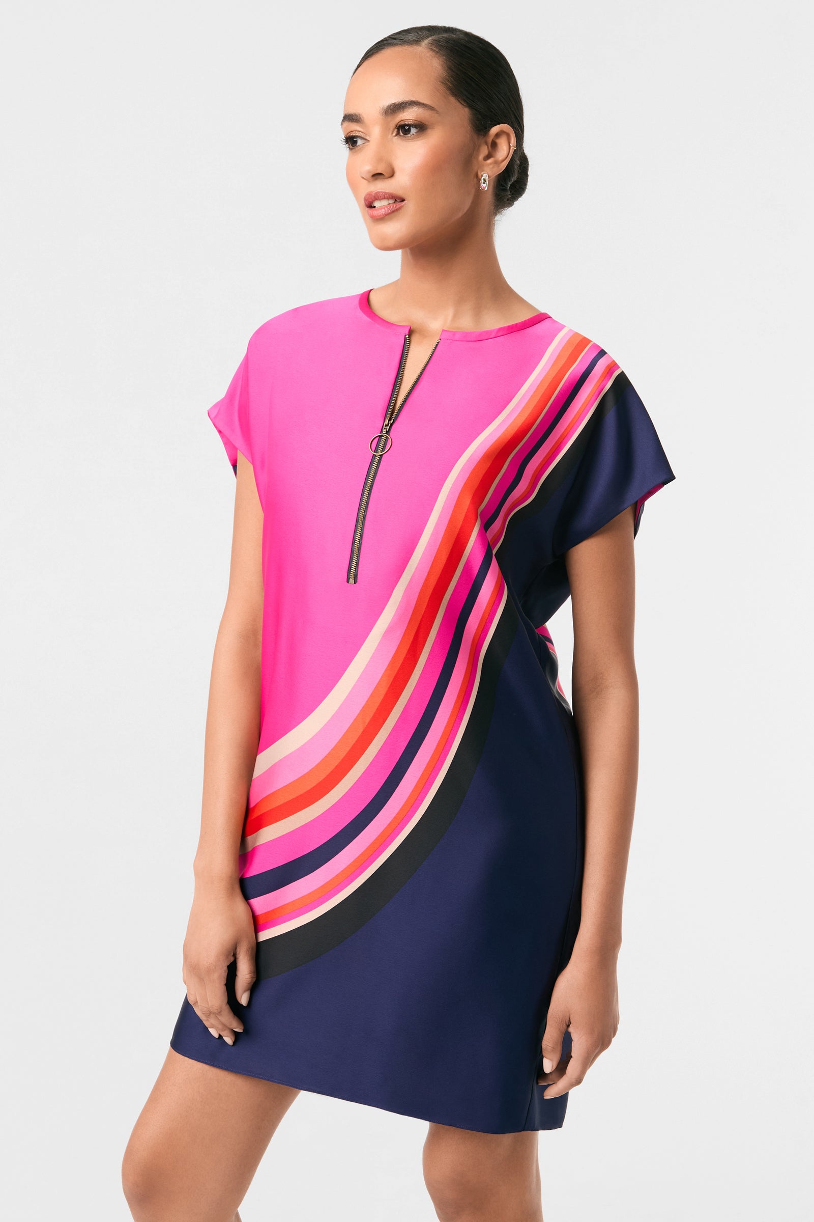 A woman models the Hasil 2 Short-Sleeve Shift Dress, which has a pink top, navy skirt, colorful curved stripes, and a front zipper. She stands against a plain light background, gazing slightly to the side.