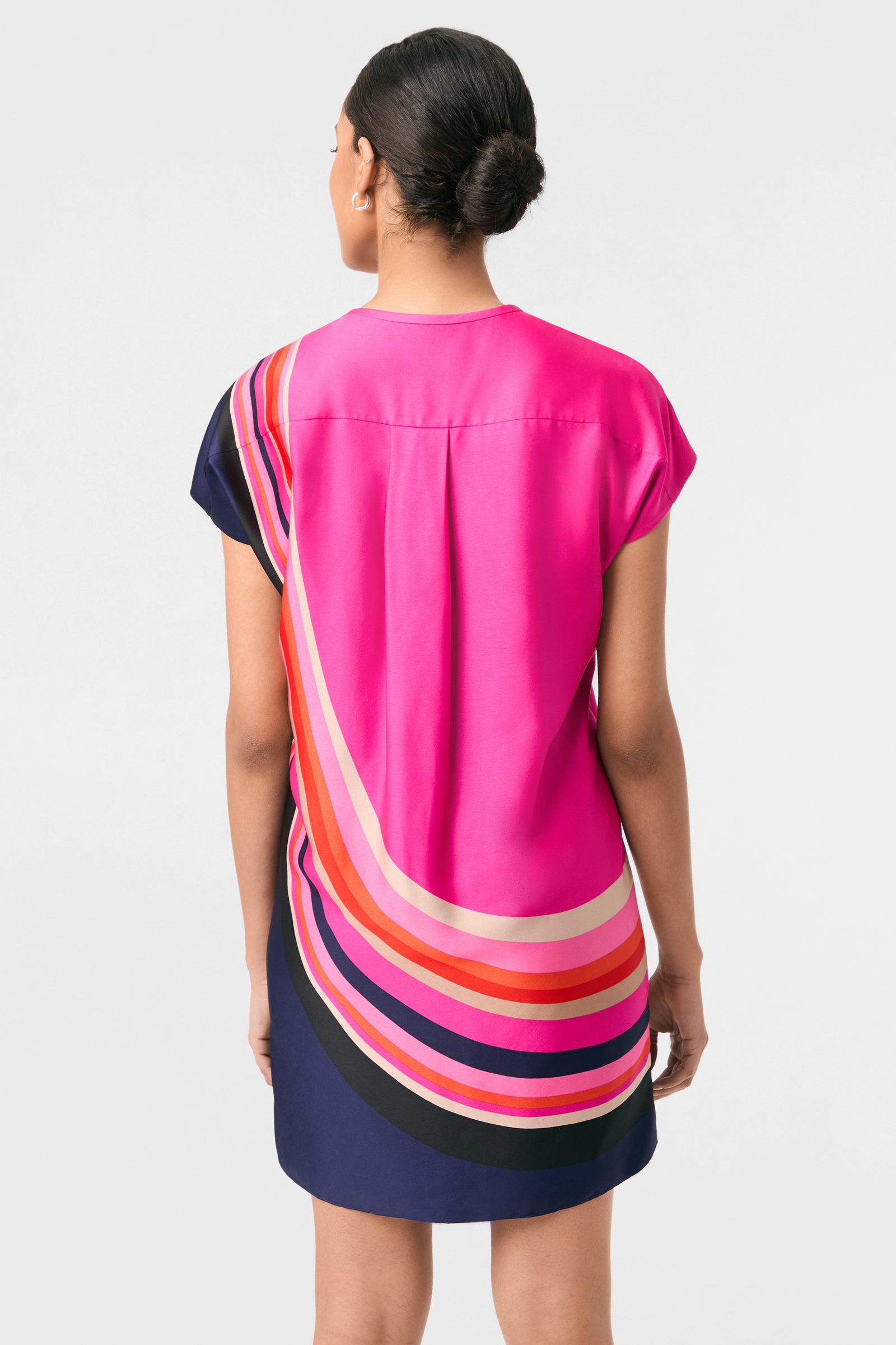 A woman with dark hair in a bun is shown from the back wearing the Hasil 2 Short-Sleeve Shift Dress, a knee-length style in bright pink with bold curved stripes of orange, cream, red, and navy against a white background.