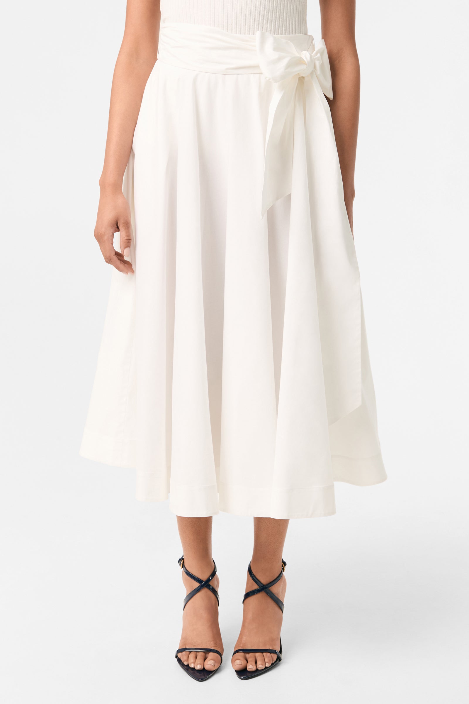 A woman wears the Amano Bow-Tie Midi Skirt in white, featuring a high waist and large bow, paired with a sleeveless white top and black strappy high-heeled sandals.