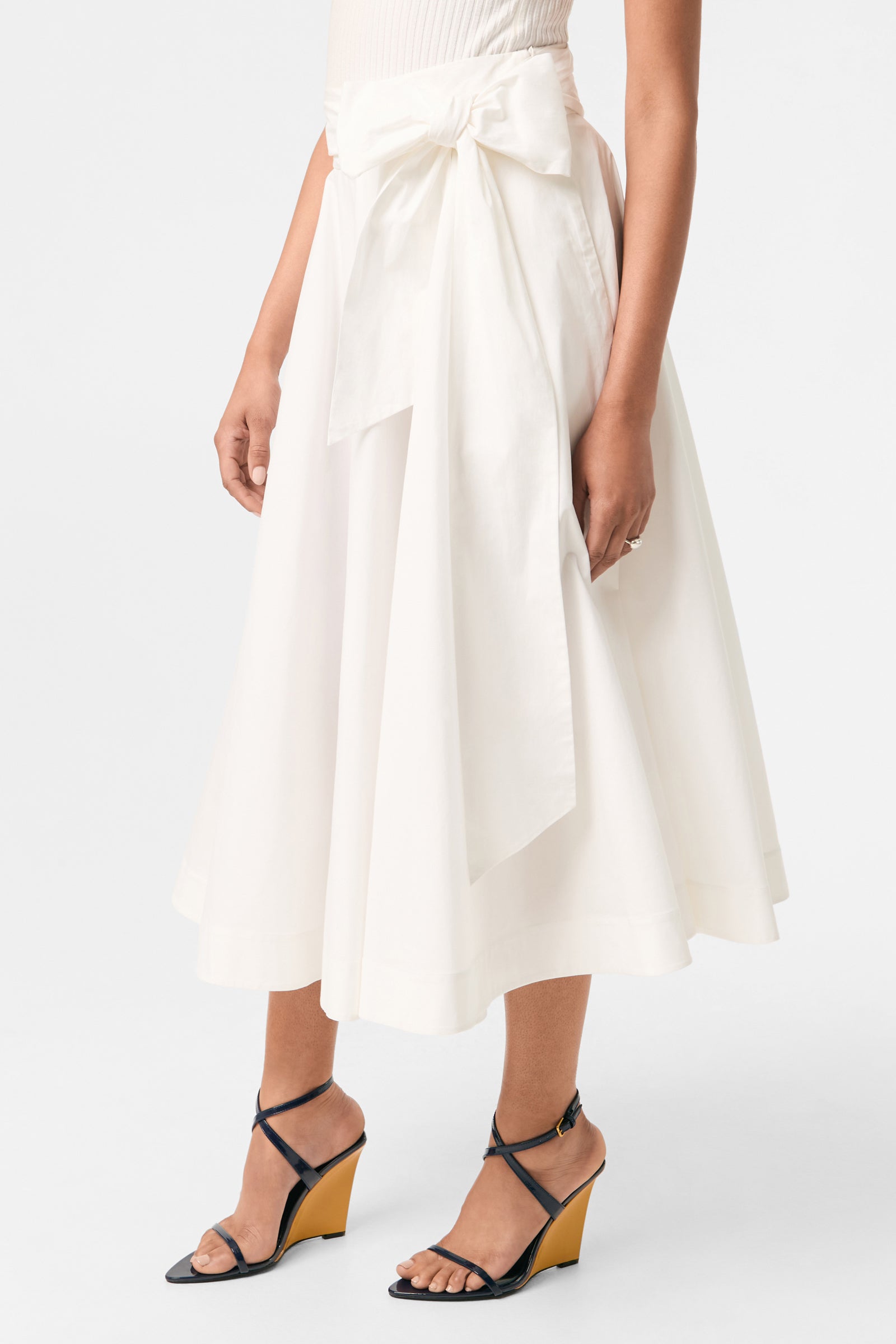 A person wears the Amano Bow-Tie Midi Skirt, a white high-waisted, midi-length skirt with a large waist bow, paired with black strappy wedge sandals against a plain white background.