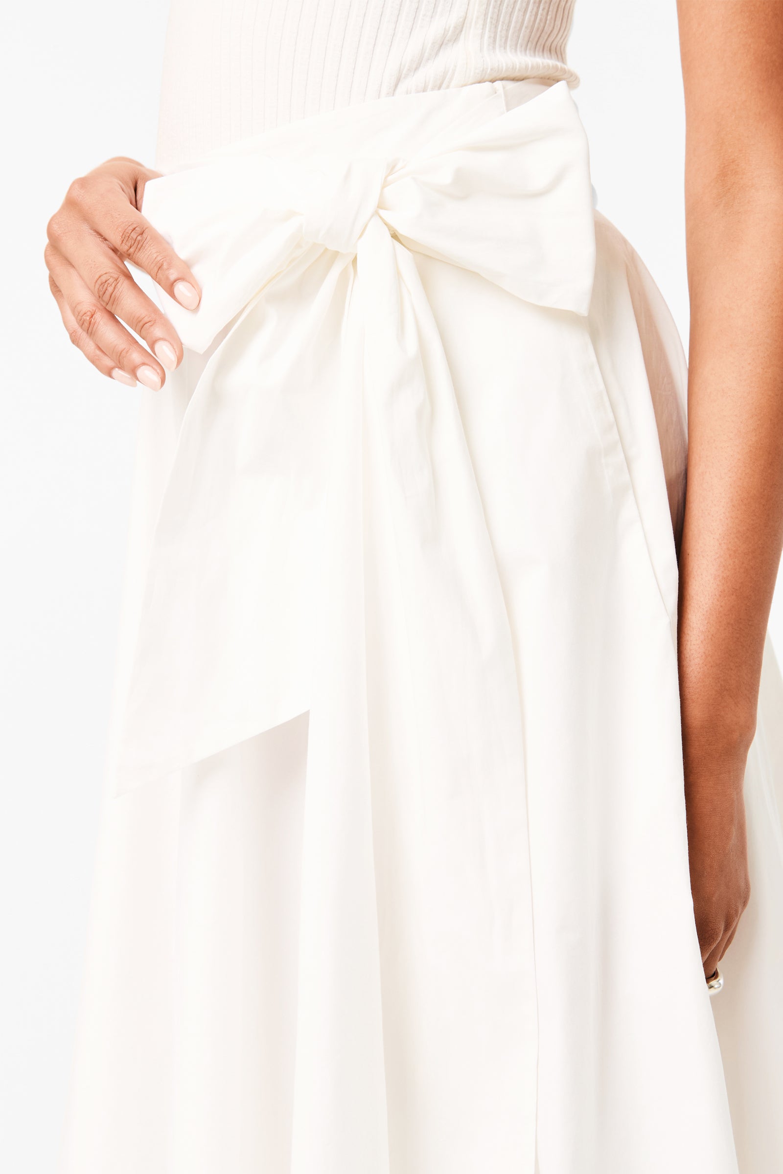A woman’s hand rests on her waist, highlighting the large bow of the Amano Bow-Tie Midi Skirt. She pairs it with a white, sleeveless top against a plain white background.