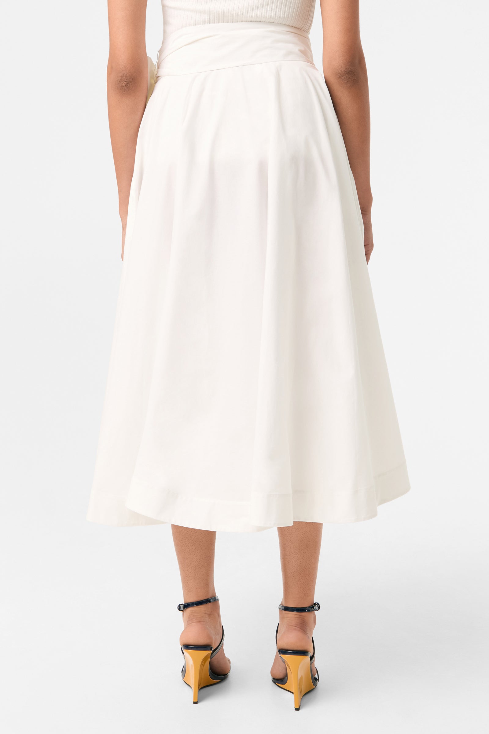 A person shown from the back against a plain white background wears the Amano Bow-Tie Midi Skirt in white, styled high-waisted and midi-length, paired with black high-heeled sandals featuring yellow accents.