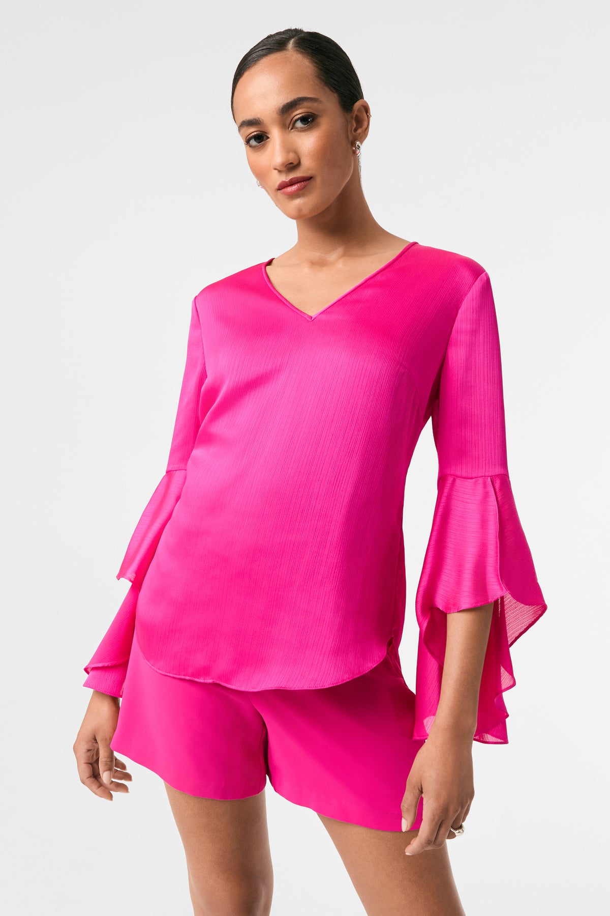 A woman stands confidently against a white background, modeling the Yuki Flounce-Sleeve Top in Trina Pink, paired with matching shorts. She faces the camera, showcasing the top’s flared, ruffled sleeves.