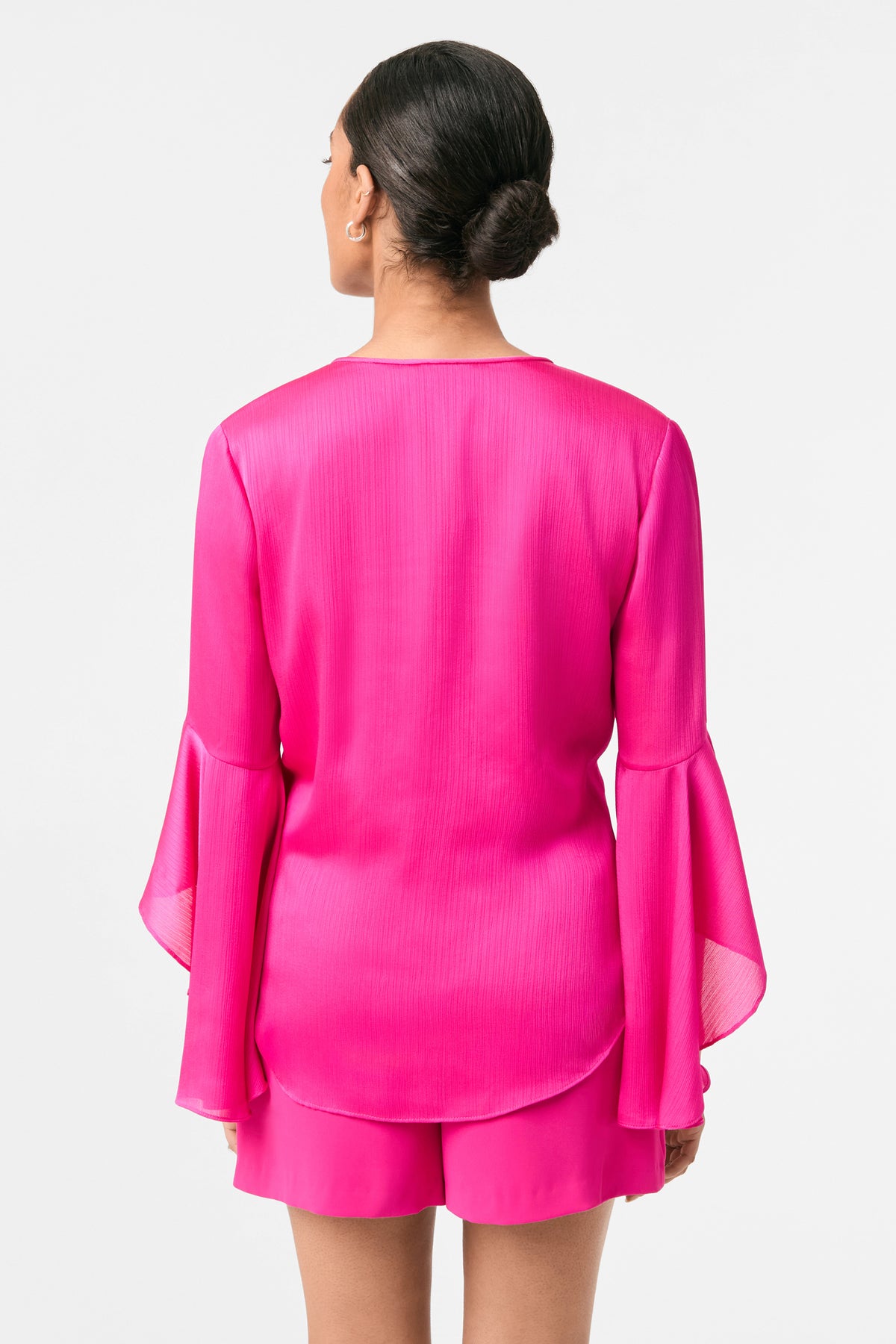 A woman with her hair in a bun is seen from behind wearing the Yuki Flounce-Sleeve Top, a bright pink long-sleeve blouse with flared cuffs, paired with matching pink shorts, set against a plain white background.