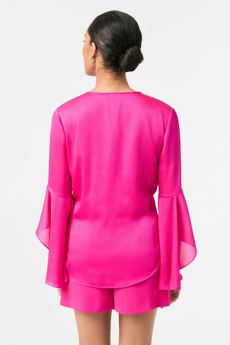 A woman with her hair in a bun is seen from behind wearing the Yuki Flounce-Sleeve Top, a bright pink long-sleeve blouse with flared cuffs, paired with matching pink shorts, set against a plain white background.