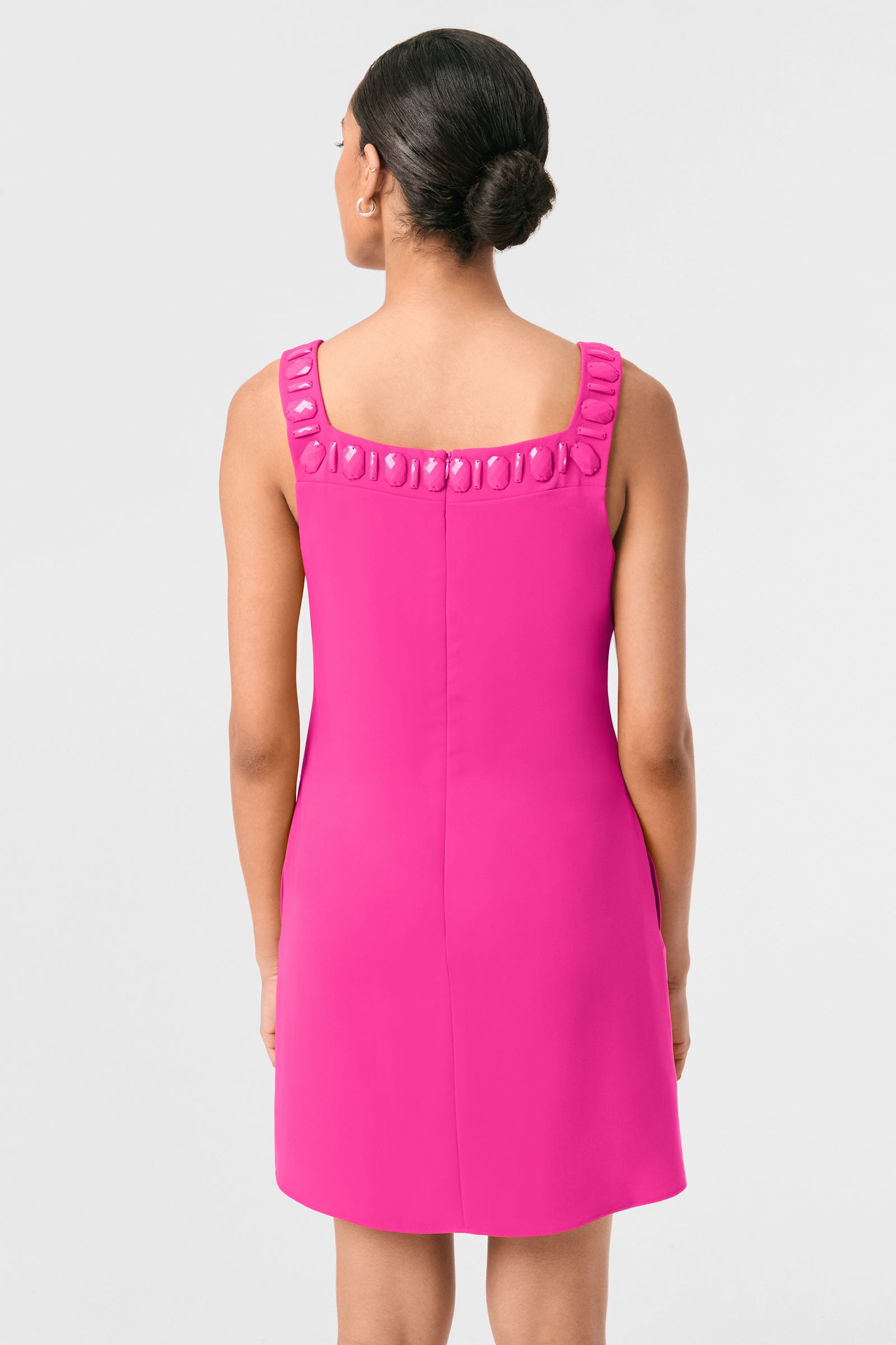 A woman with her hair in a neat bun stands facing away, wearing the Yokuna Jewel Neck Mini Dress—a sleeveless, bright pink mini dress featuring a square neckline adorned with decorative embellishments—against a plain light gray background.